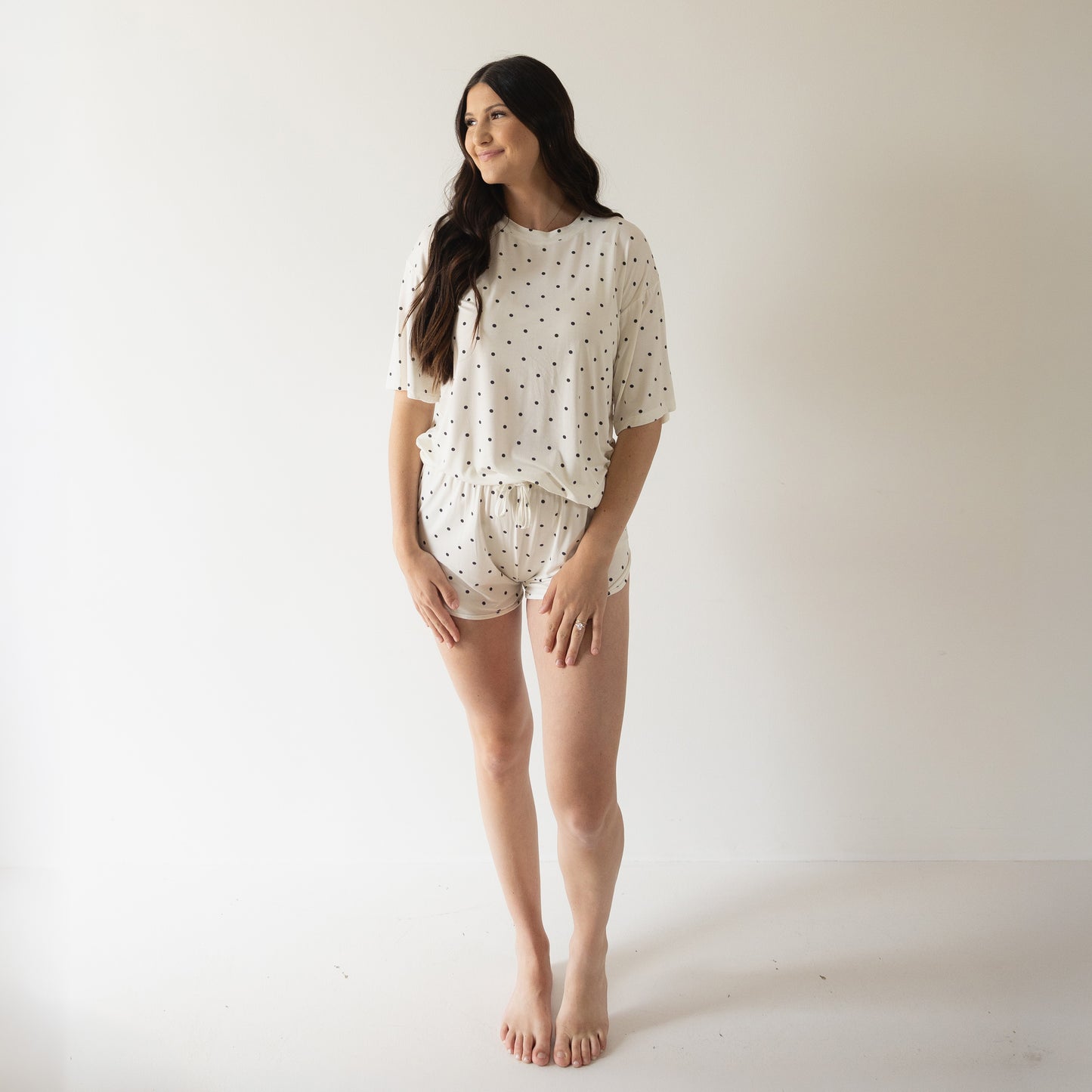 A woman stands barefoot in a minimalist room, wearing the Women's Bamboo Short Set | Annie by forever french baby with black polka dots. She has long dark hair and smiles slightly while looking to the side.