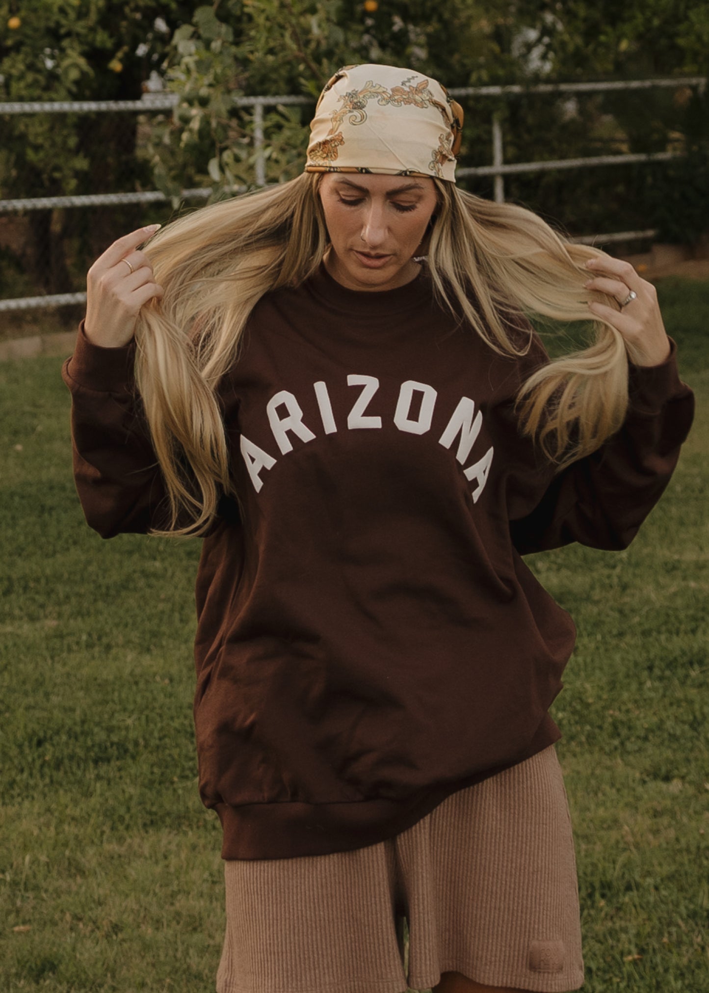 A woman wears the forever french baby Adult Sweatshirt | ff Arizona in brown, paired with light brown shorts and a patterned bandana, standing on grass with trees and a fence behind her, holding her long blonde hair.