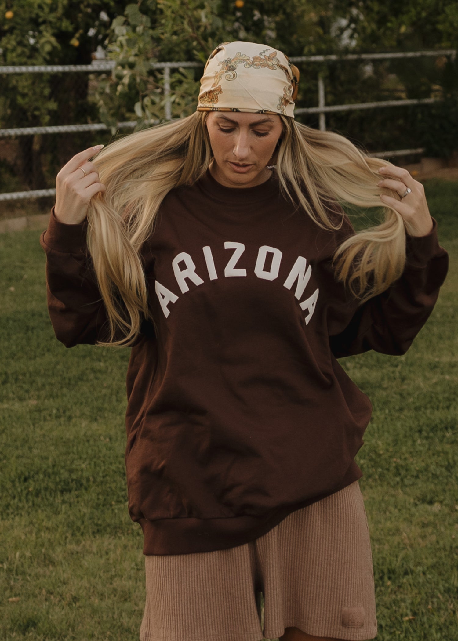 A woman wears the forever french baby Adult Sweatshirt | ff Arizona in brown, paired with light brown shorts and a patterned bandana, standing on grass with trees and a fence behind her, holding her long blonde hair.