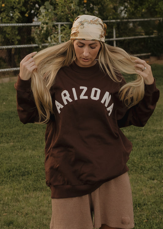 A woman wears the forever french baby Adult Sweatshirt | ff Arizona in brown, paired with light brown shorts and a patterned bandana, standing on grass with trees and a fence behind her, holding her long blonde hair.