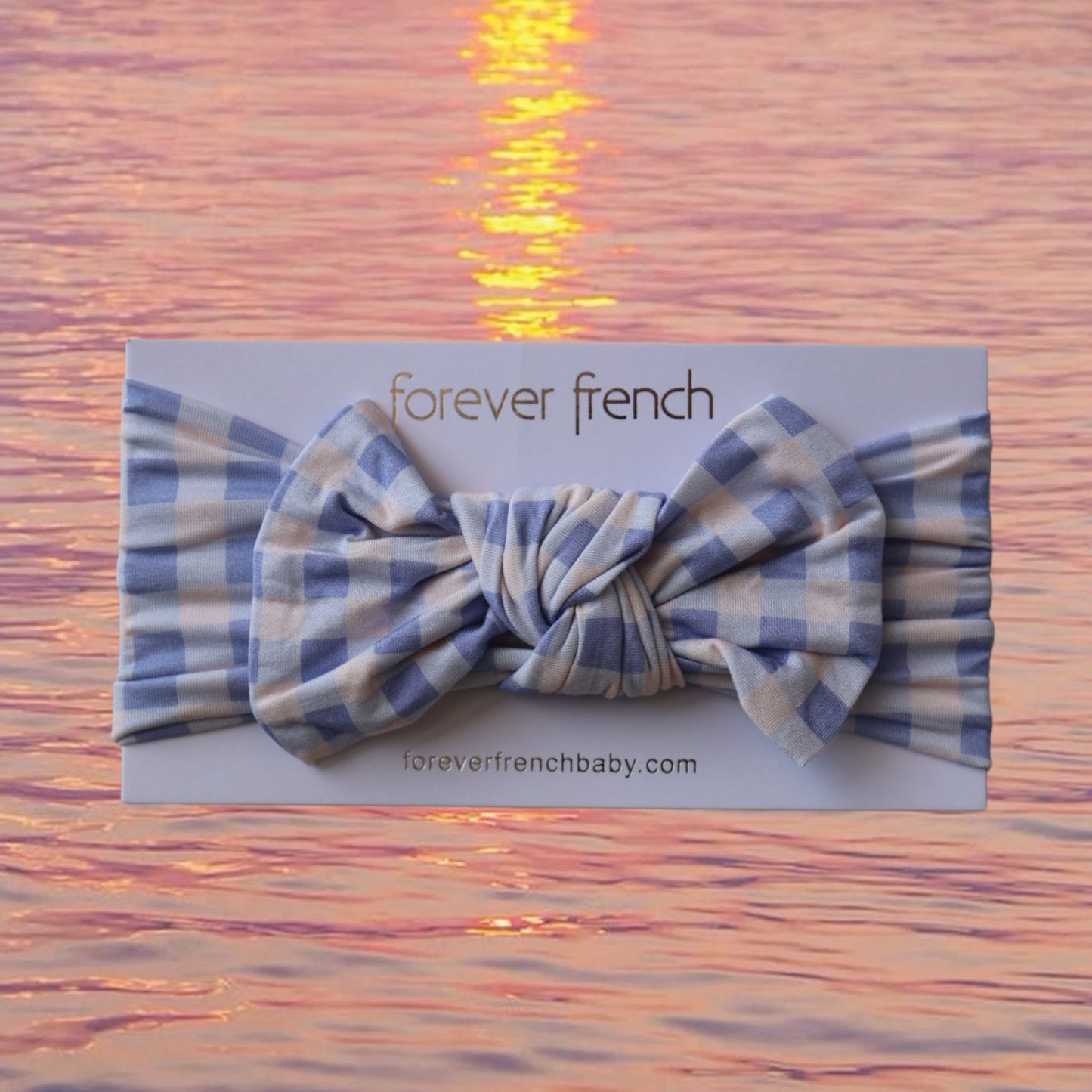 A Bamboo Head Wrap | Marilyn by forever french baby, featuring a blue and white checkered bow, displayed on a branded card with a pink sunset over water in the background.