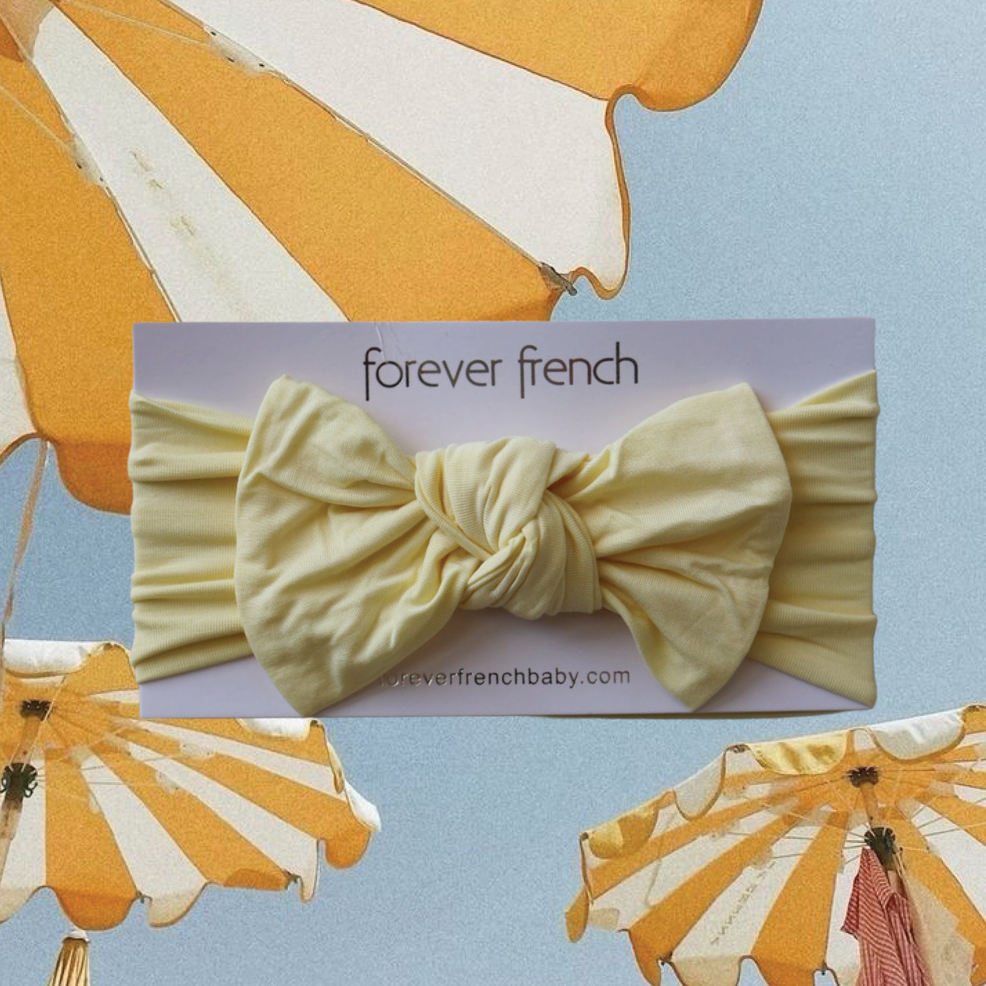 A Bamboo Head Wrap in the shade “Sunny” by forever french baby is displayed on a card labeled with the brand’s name, set against a background of yellow-and-white striped umbrellas and sky.