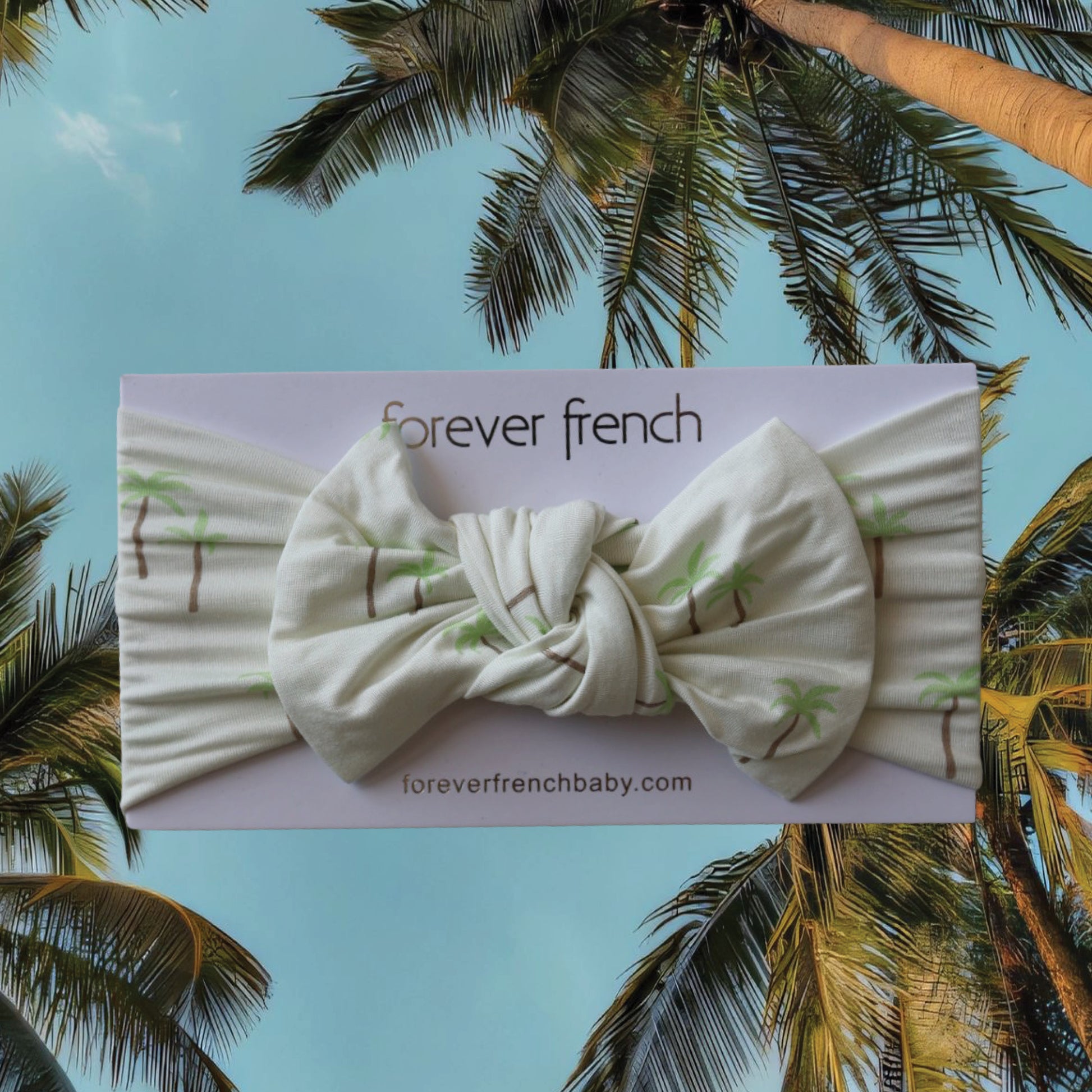 A soft bamboo head wrap from forever french baby, called the Bamboo Head Wrap | Palm, features a white fabric with a green palm print and an oversized bow. Shown against a backdrop of sky and palm trees, this playful accessory is perfect for little ones and adds a charming touch to any outfit.