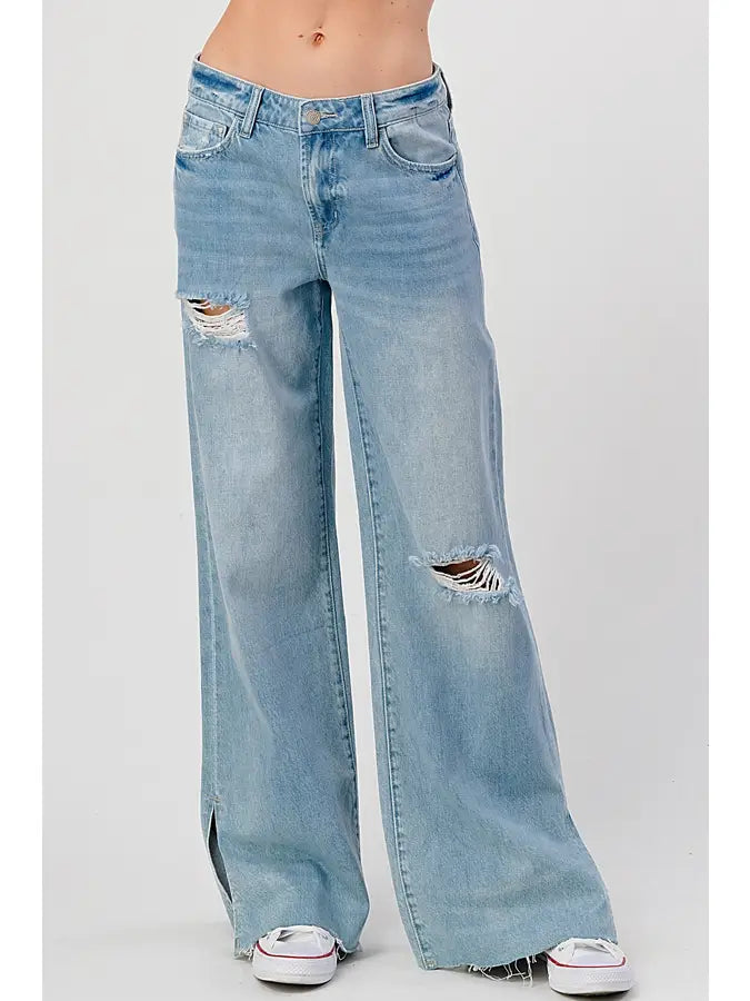 Someone dressed in medium wash High Rise Skater Jeans from I&M JEAN, INC. pairs them with white sneakers. These women's jeans showcase distressed accents on the left knee and right thigh, a frayed hem, and front pockets, adding a stylish contrast against the simple background.