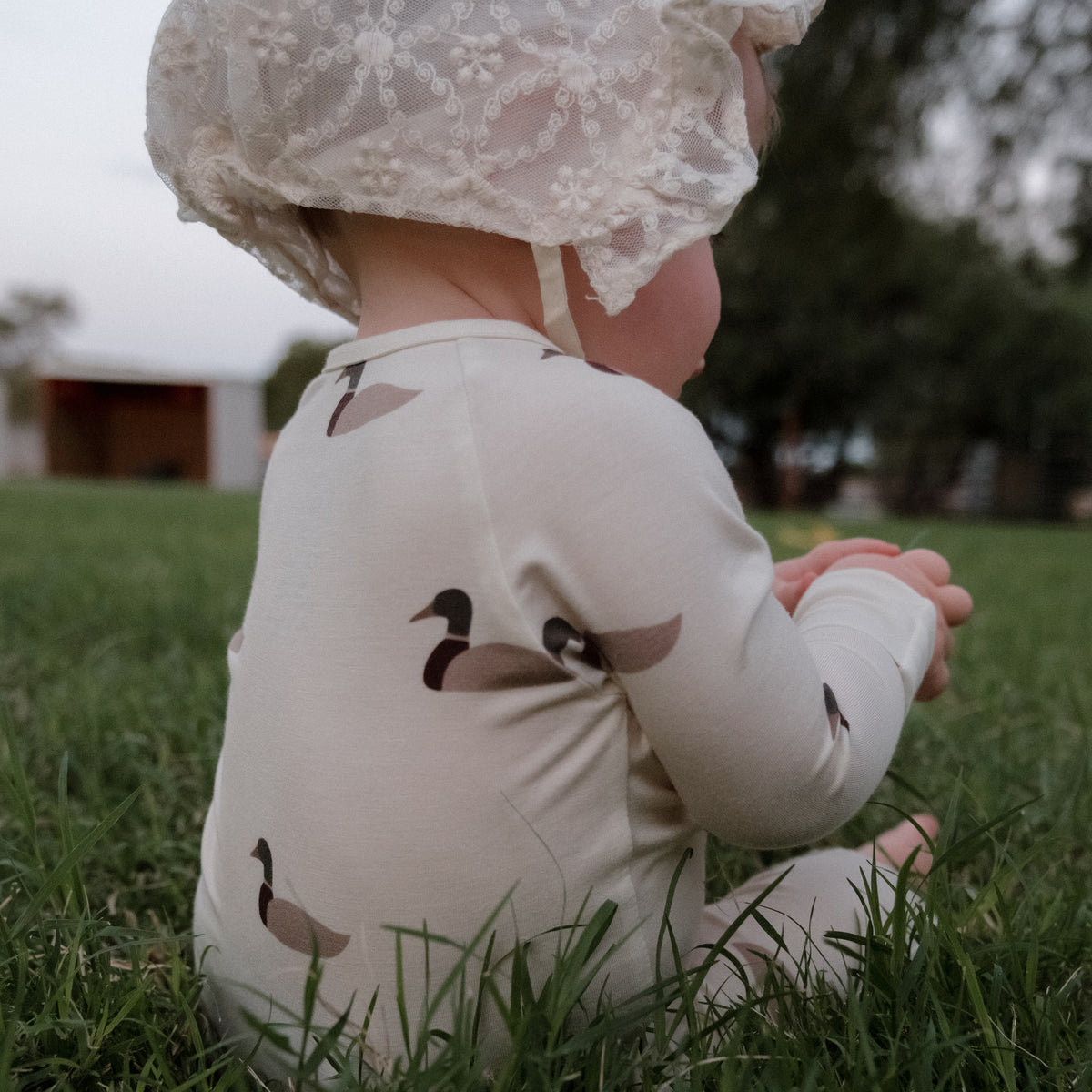A baby wearing forever french baby’s Bamboo Zip Pajamas in the Mallard print, made from hypoallergenic fabric and featuring adorable ducks, sits on grass outdoors with their back to the camera, topped with a lace bonnet.