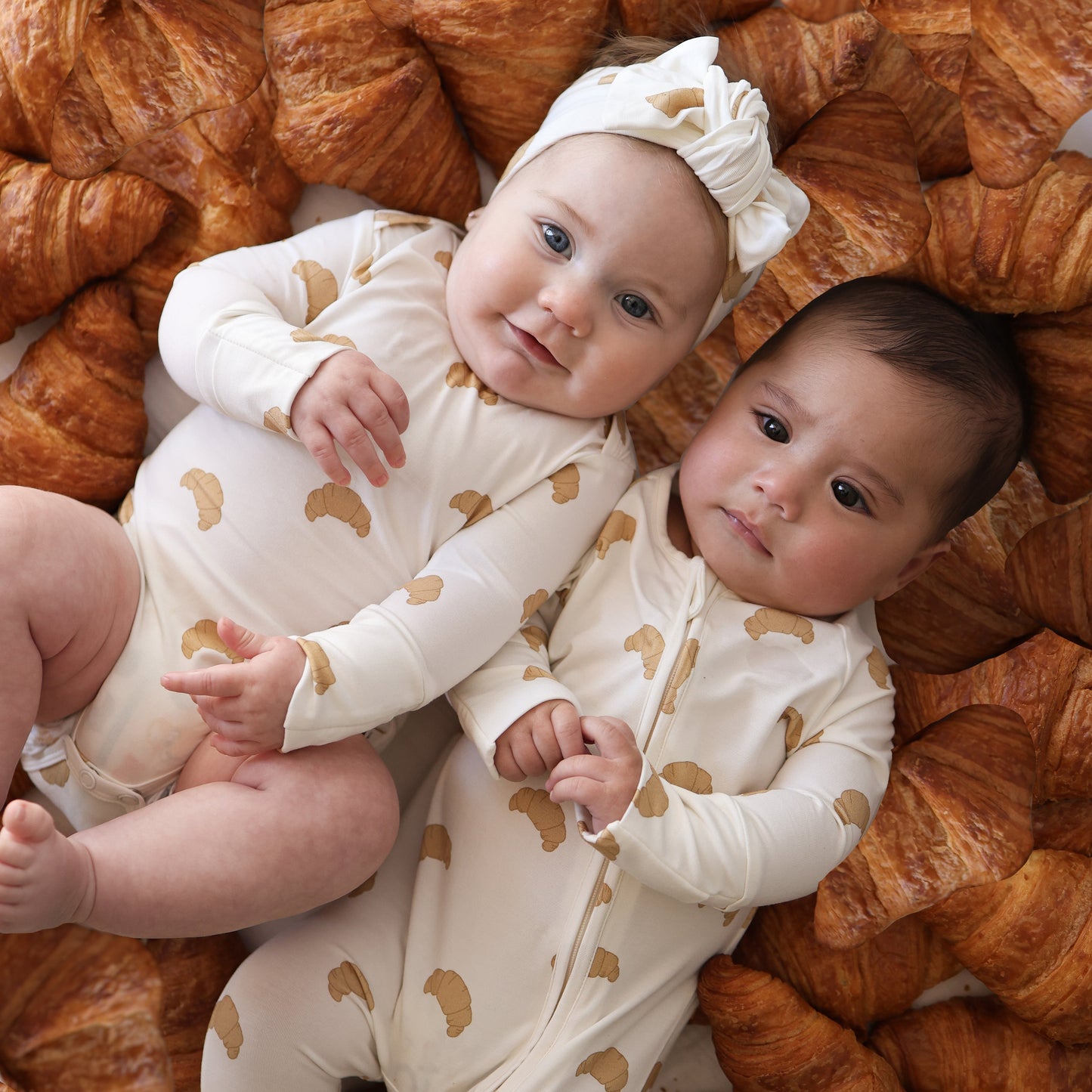 Two babies in matching forever french baby Bamboo Zip Pajamas | Croissants lie side by side on a croissant bed. One wears a bow headband, and both look content in their soft, hypoallergenic bamboo baby clothing.