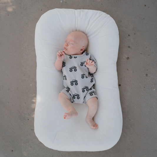 A sleeping baby wears the forever french baby French Terry Romper | Monster Truck, a gray newborn short sleeve romper with black monster truck prints, while lying on a white cushioned pad atop a light-colored surface.