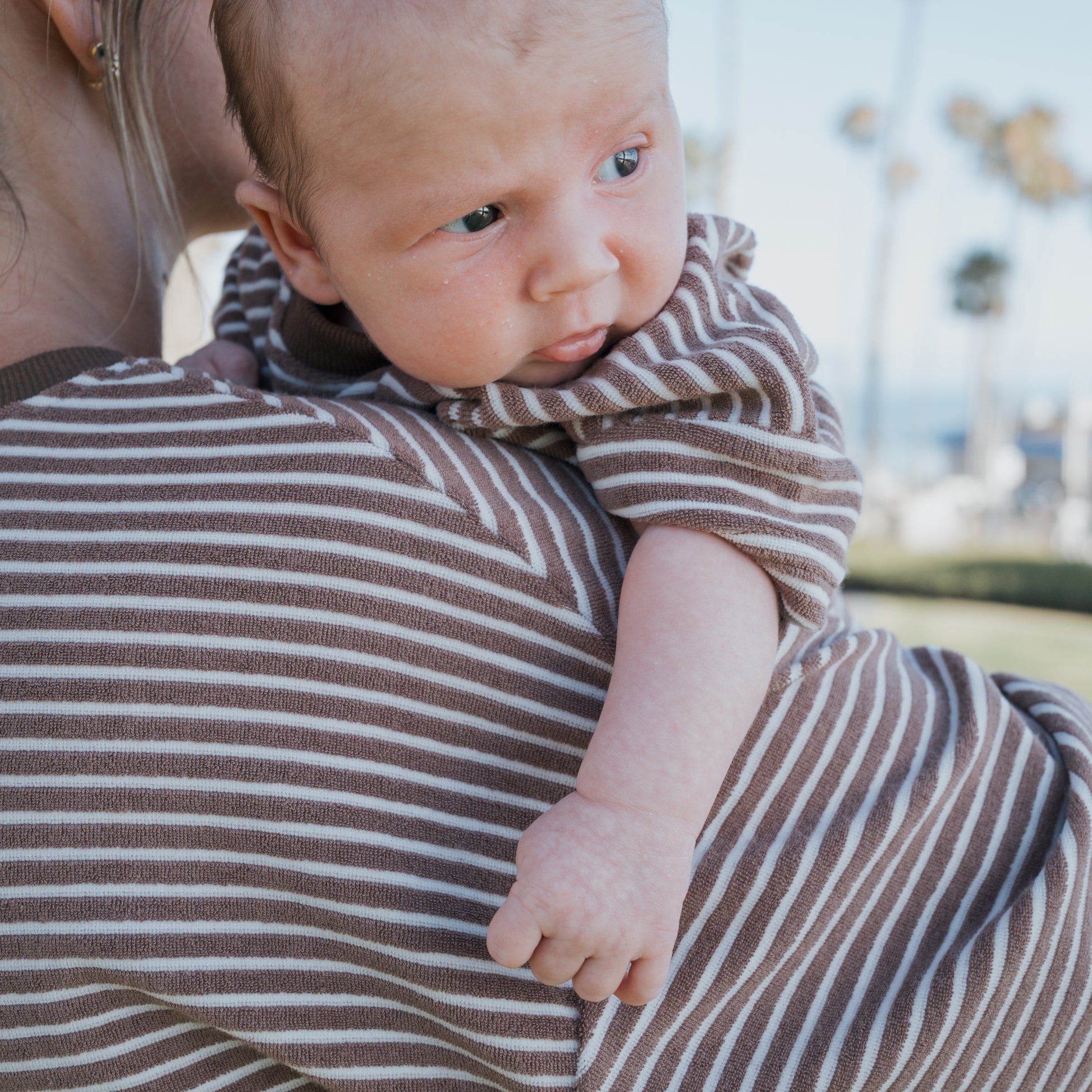 A baby wears the forever french baby Terry T-Shirt Romper in Cola Stripe, held on someone's shoulder in matching outfits. The curious baby looks to the side, with blurred palm trees softly framing the outdoor scene.
