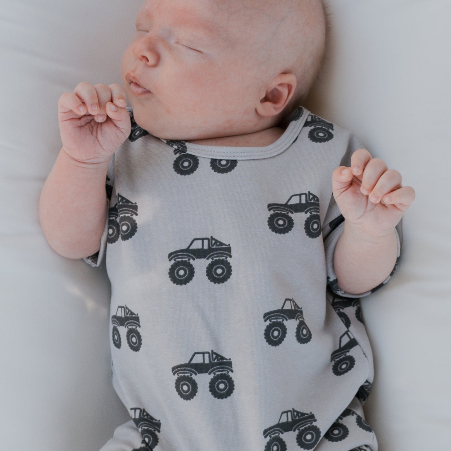 A baby sleeps on a white surface wearing the forever french baby French Terry Romper | Monster Truck—a light gray, short-sleeve romper with black monster trucks that keeps them comfy, hands relaxed and slightly raised near their face.
