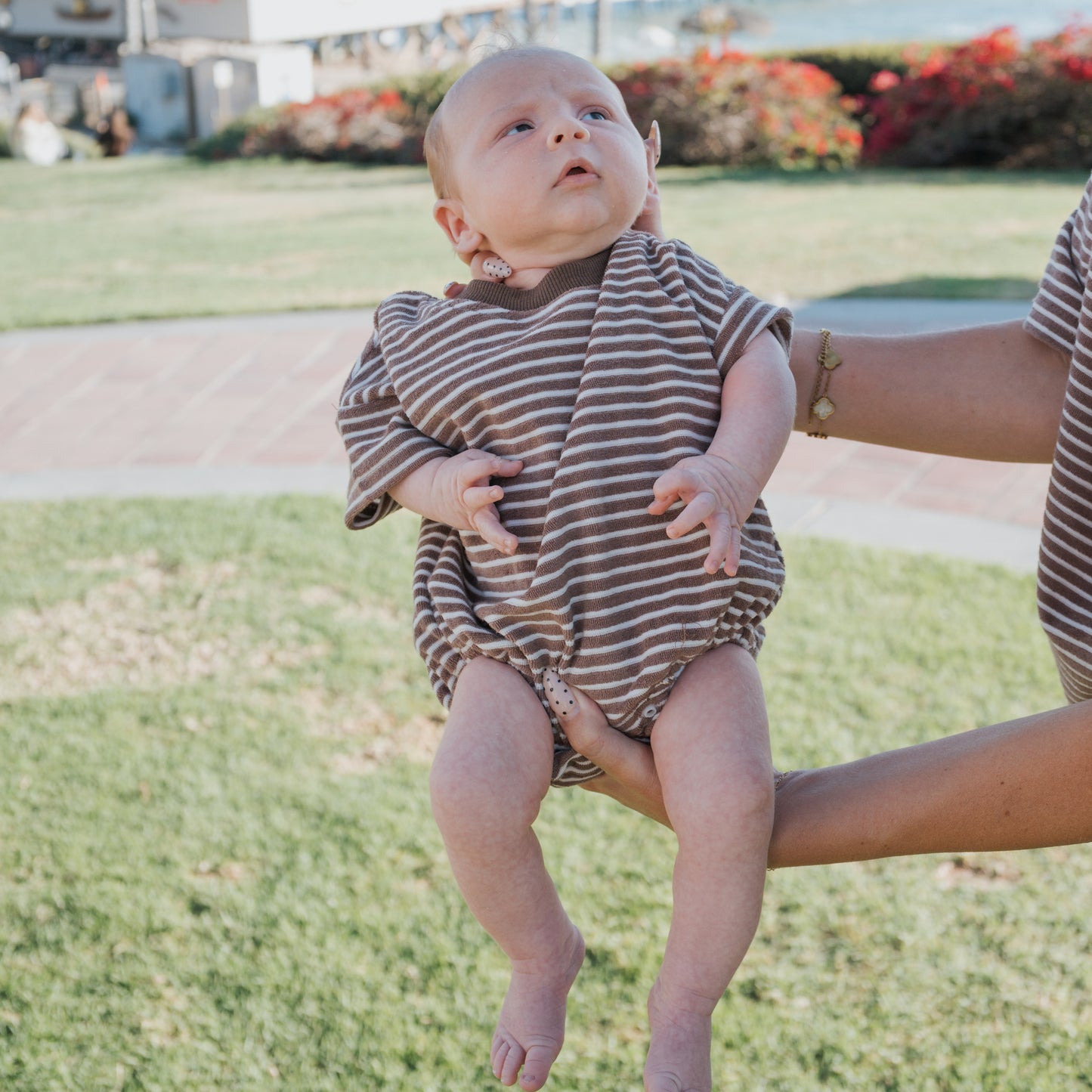 A baby wears the forever french baby Terry T-Shirt Romper in Cola Stripe while being held outdoors by an adult, with grass, flowers, and buildings visible in the bright daylight; only the adult’s arms and torso appear.