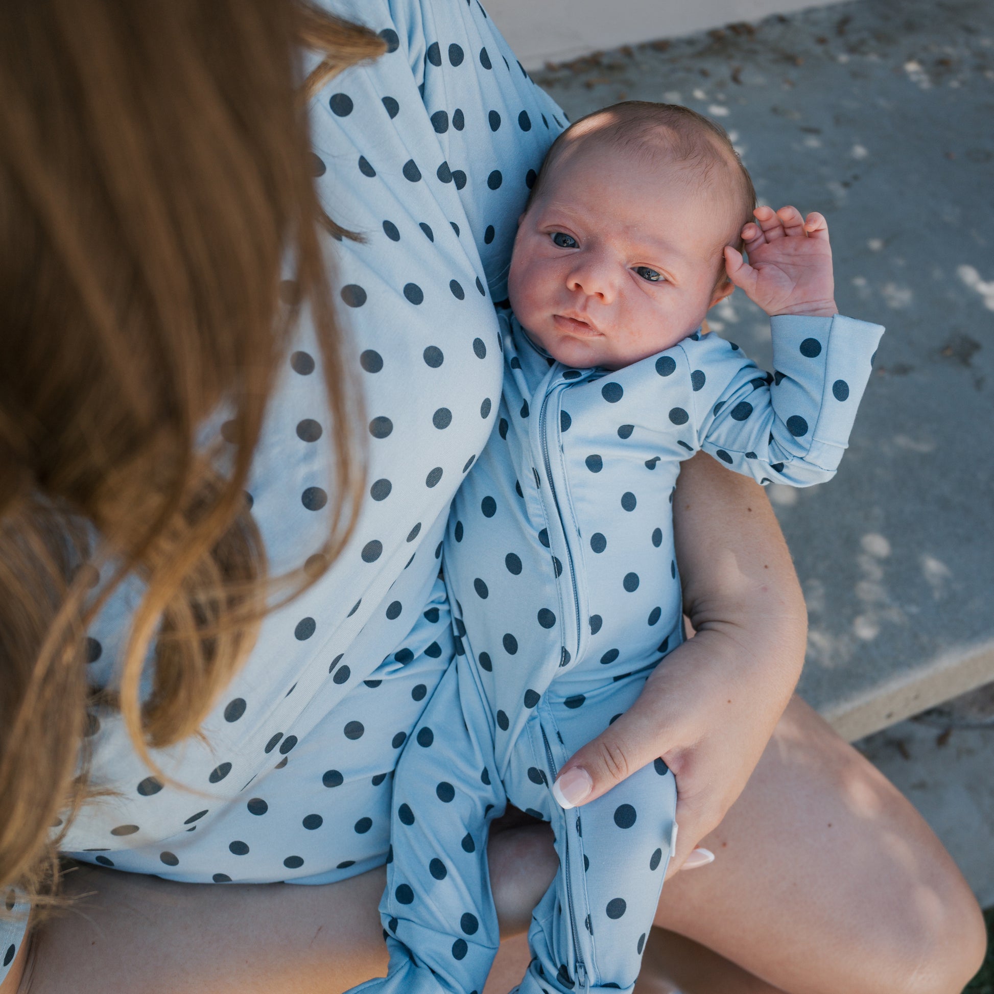 A baby wearing the forever french baby Bamboo Zip Pajamas | Sully rests in an adult's arms. Both are dressed in matching blue polka dot outfits, with the little one cozy in breathable sleepwear. They are sitting together on a concrete step outdoors.