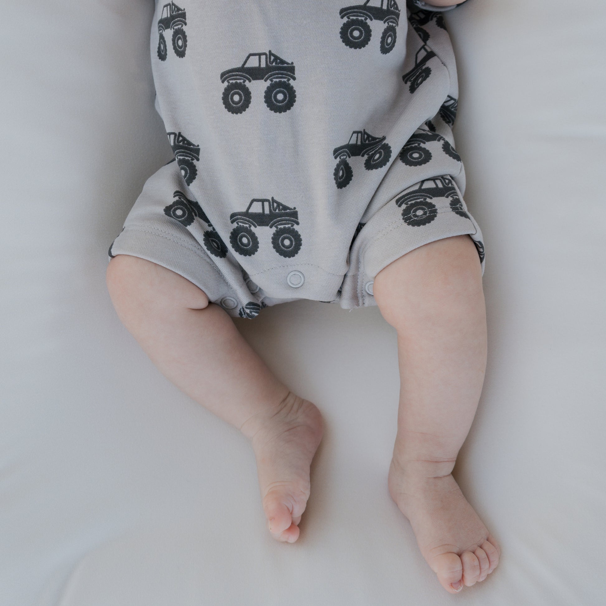 A baby’s legs and lower torso are shown while lying on a white surface, dressed in the forever french baby French Terry Romper | Monster Truck, a breathable light gray romper featuring a black monster truck pattern.