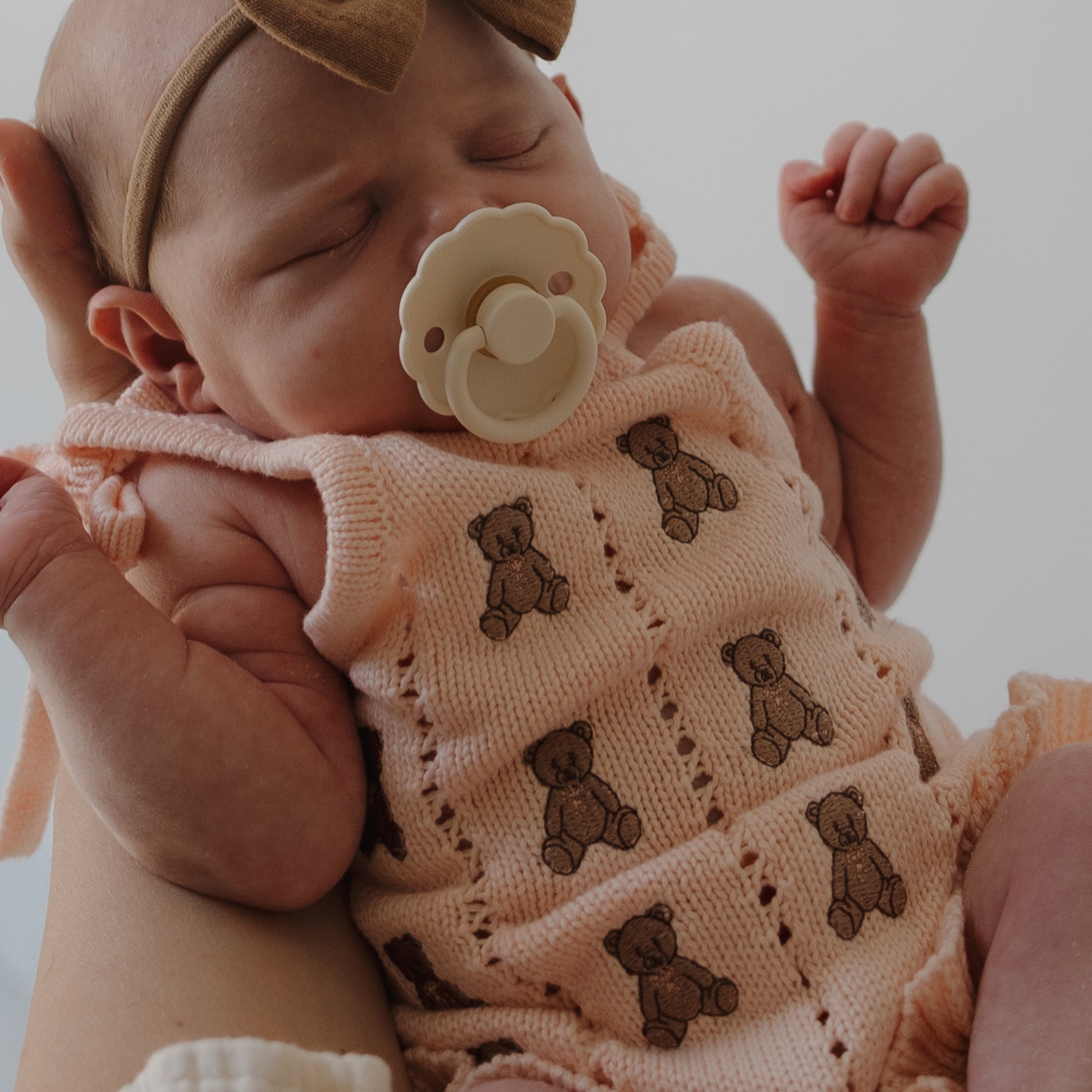 A newborn with a pacifier and brown bow headband is cradled in an adult's arm, wearing the forever french baby Knit Ruffle Romper | Lottie, a peach cotton outfit featuring embroidered teddy bear patterns.