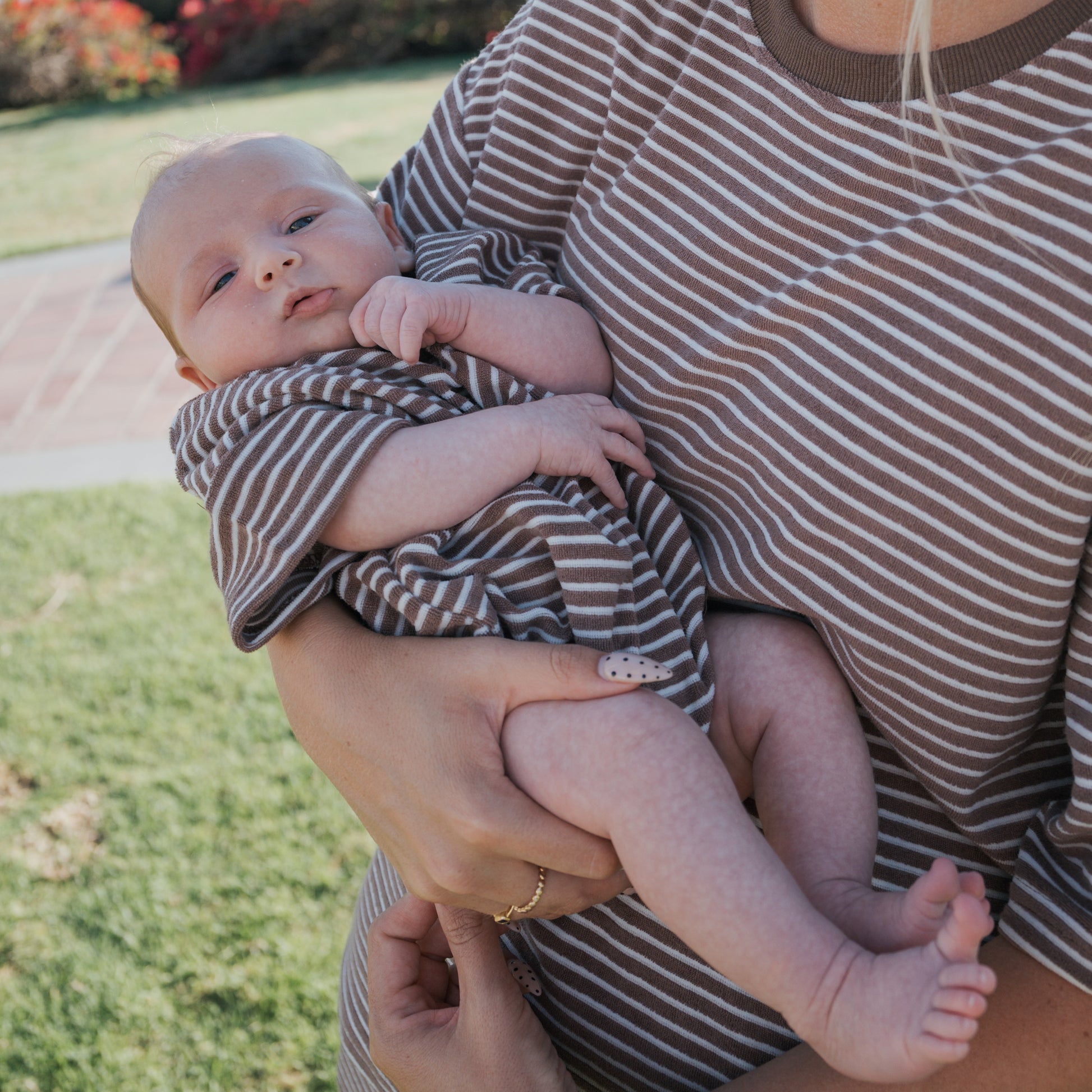 A baby wears the forever french baby Terry T-Shirt Romper in Cola Stripe, cradled in an adult's arms. Both are dressed in matching family outfits outdoors on grass, with bushes and a sidewalk behind them.