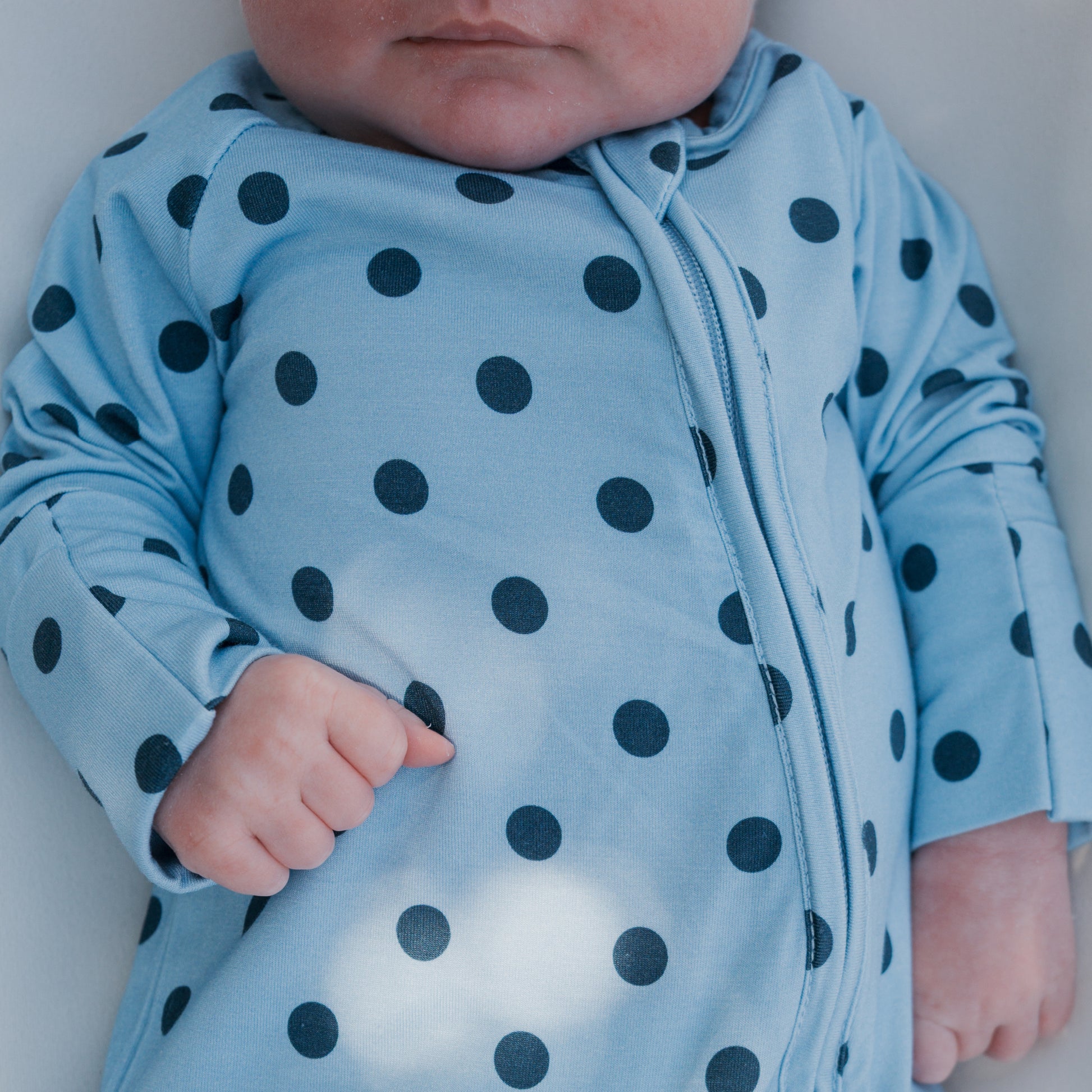 A baby wearing the forever french baby Bamboo Zip Pajamas | Sully, a light blue breathable sleepwear with black polka dots, lies on a white surface with only the lower face and body visible, ensuring comfort throughout the night.