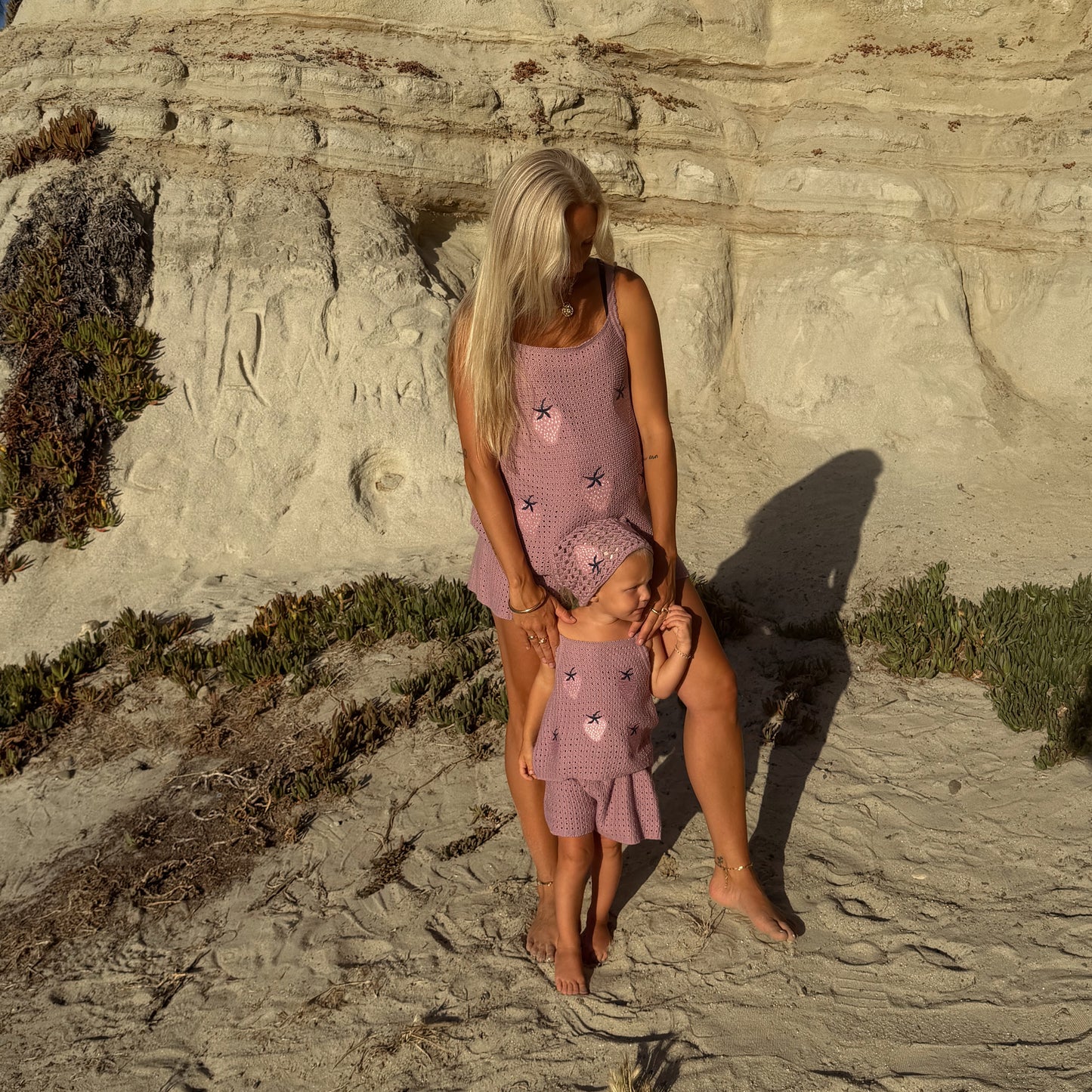 A woman and a young girl stand barefoot on sandy ground near rocky cliffs, sunlight and plants casting soft shadows as they wear the matching Matilda Women's Knit Short Set by forever french baby, crafted from soft knit fabric for ultimate comfort.