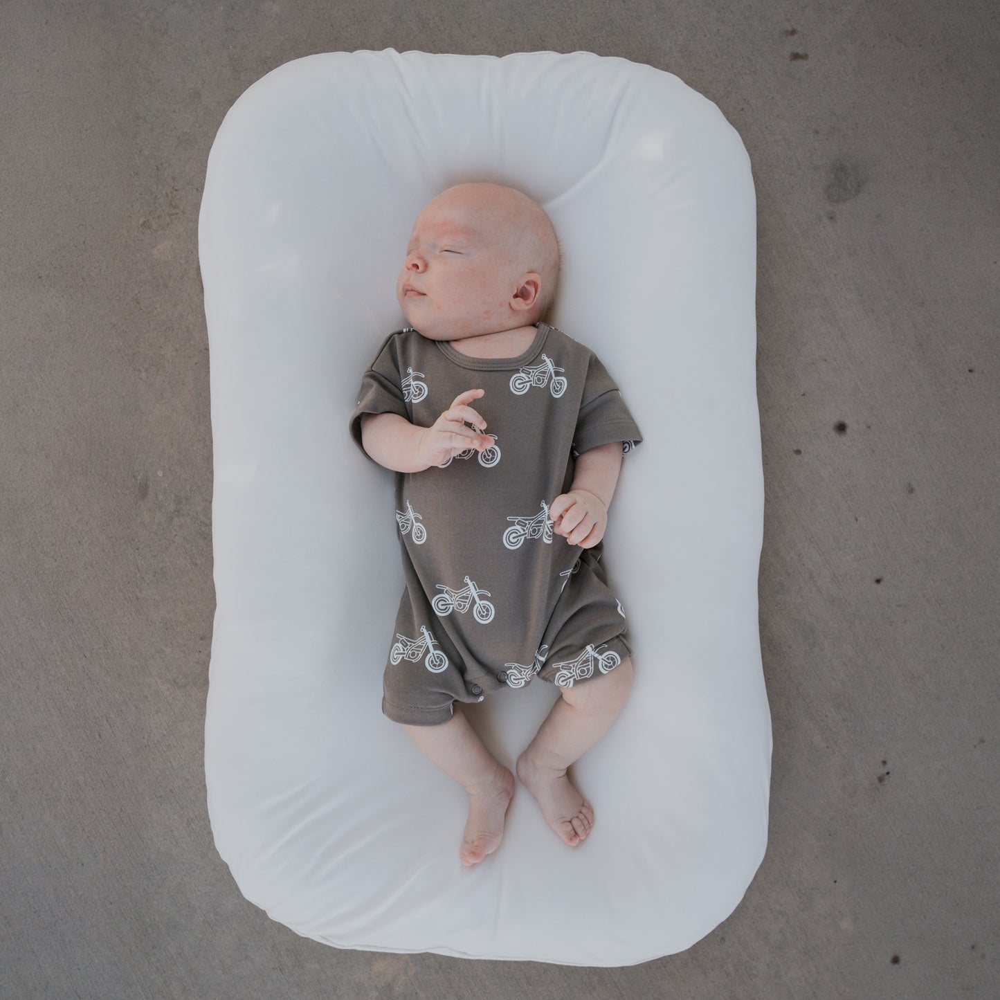 A newborn wearing the forever french baby French Terry Romper in the Dirt Bike print lies on a white cushioned pad atop a gray concrete floor.
