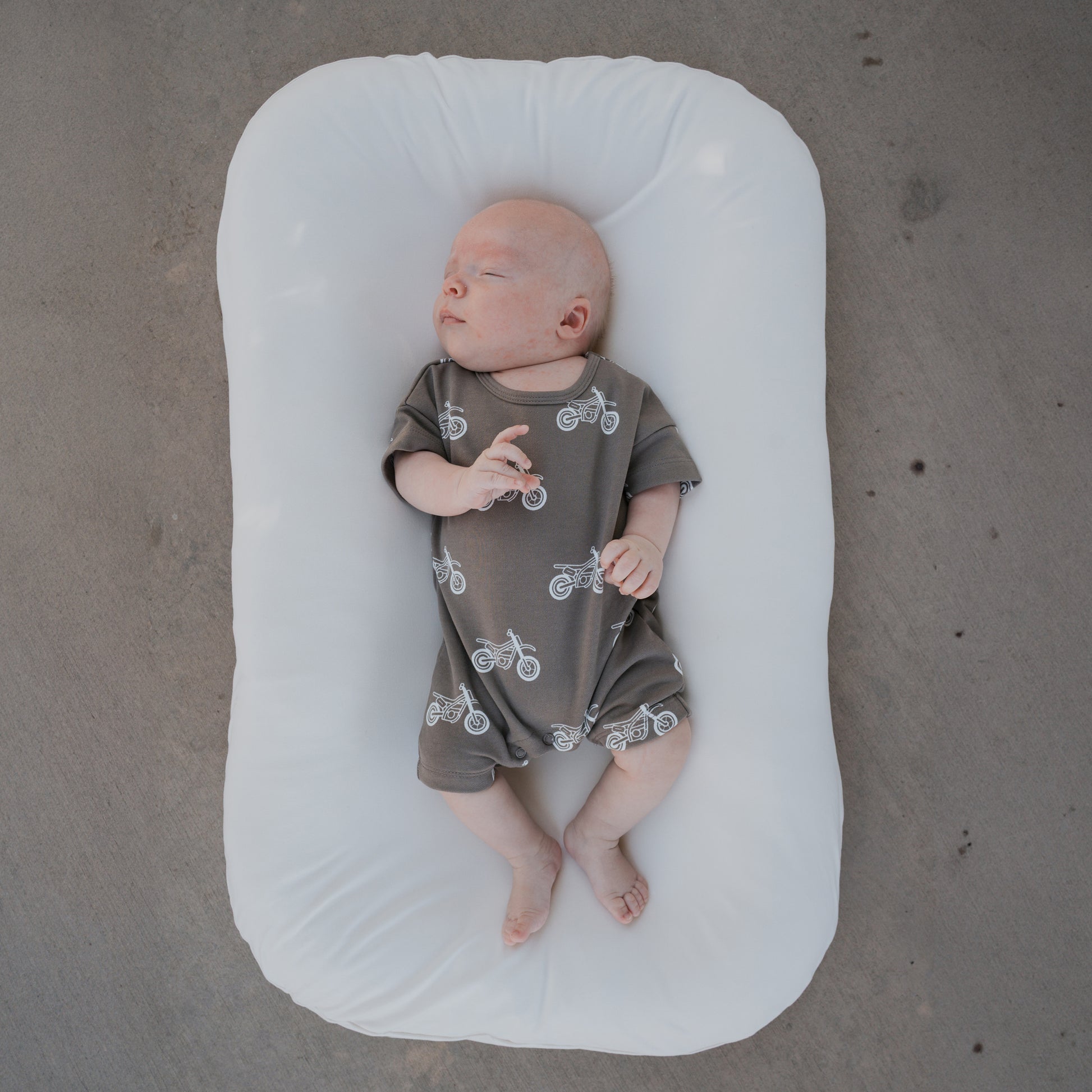 A newborn wearing the forever french baby French Terry Romper in the Dirt Bike print lies on a white cushioned pad atop a gray concrete floor.