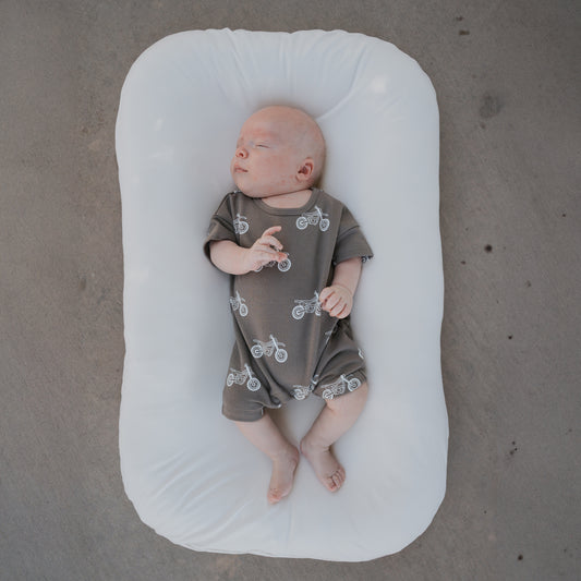 A newborn wearing the forever french baby French Terry Romper in the Dirt Bike print lies on a white cushioned pad atop a gray concrete floor.