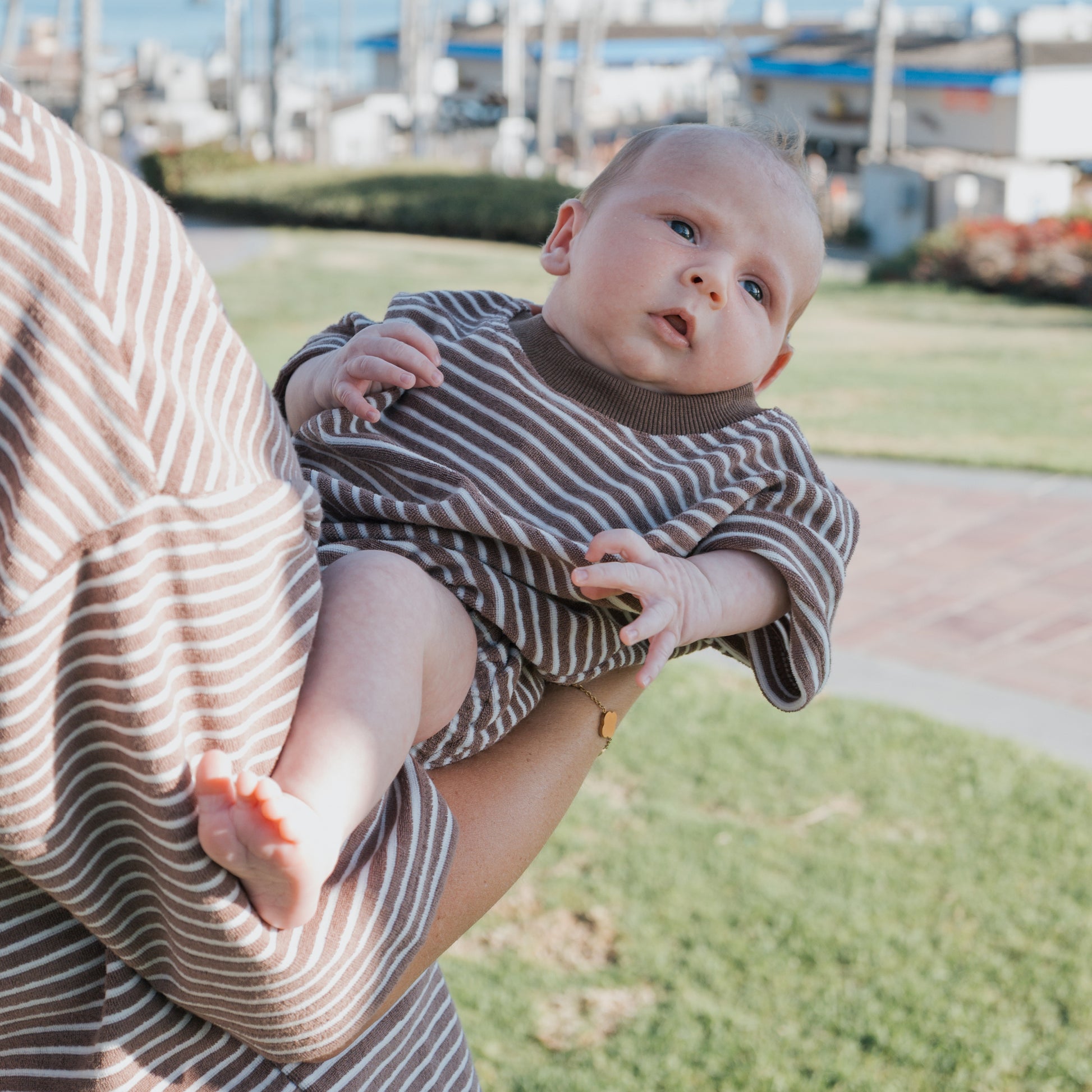 A baby wears the forever french baby Terry T-Shirt Romper in Cola Stripe, held outdoors by an adult in a matching striped top, both in coordinated family outfits with grass and blurred buildings in the background.