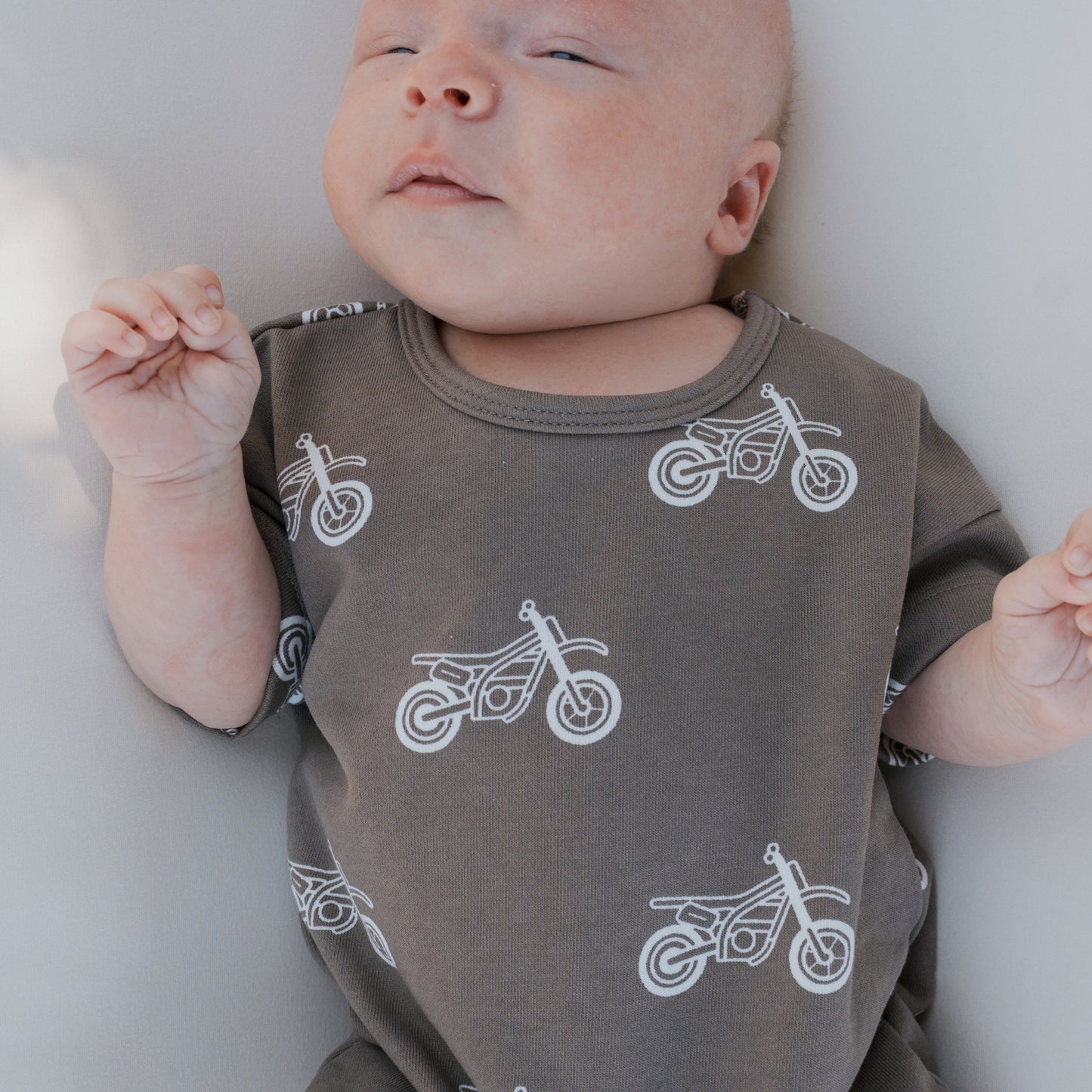 A baby in the forever french baby French Terry Romper | Dirt Bike—a dark gray cotton outfit with white motorcycle prints—lies on a light surface, eyes half-closed and hands up near the face; a cute pick for newborns.