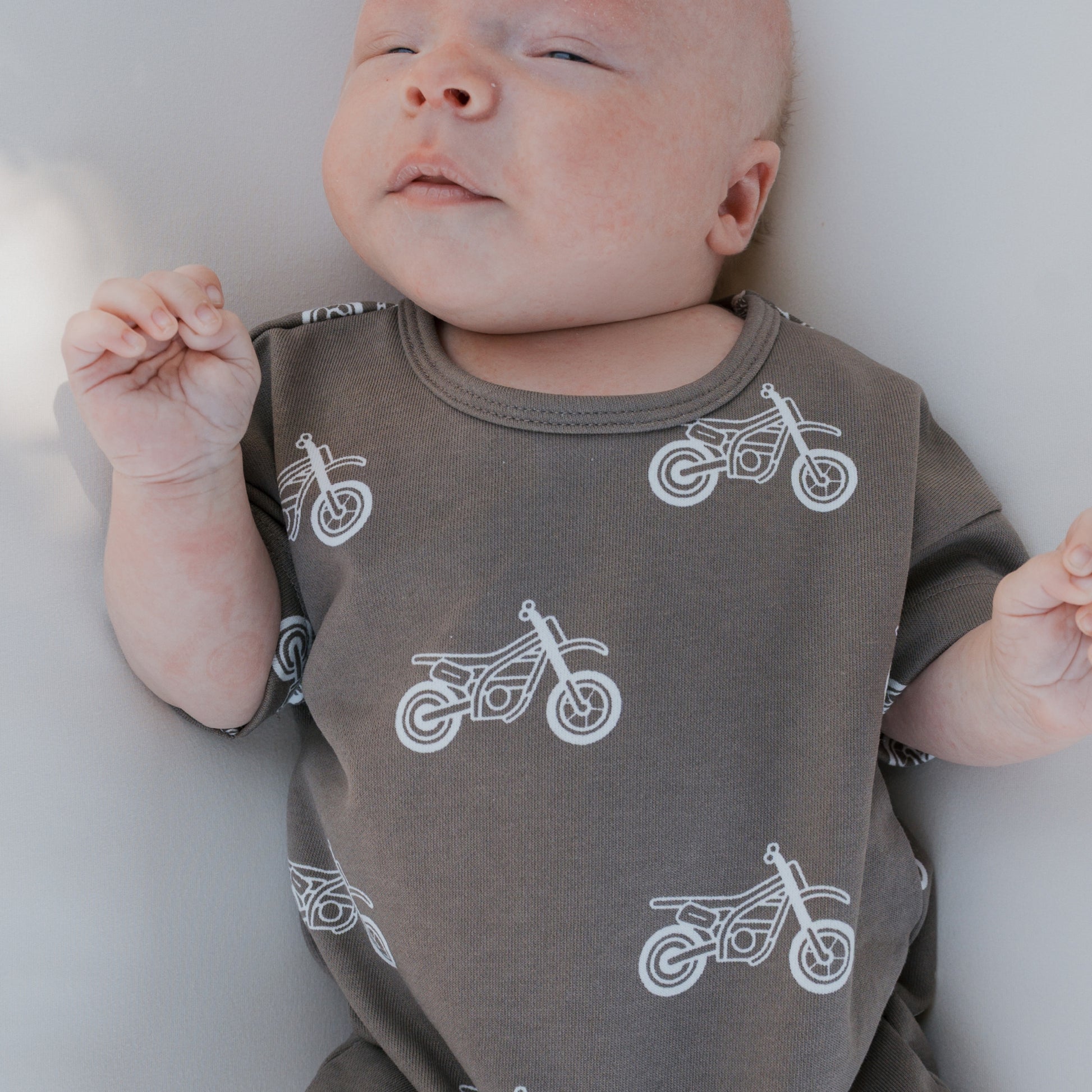 A baby in the forever french baby French Terry Romper | Dirt Bike—a dark gray cotton outfit with white motorcycle prints—lies on a light surface, eyes half-closed and hands up near the face; a cute pick for newborns.