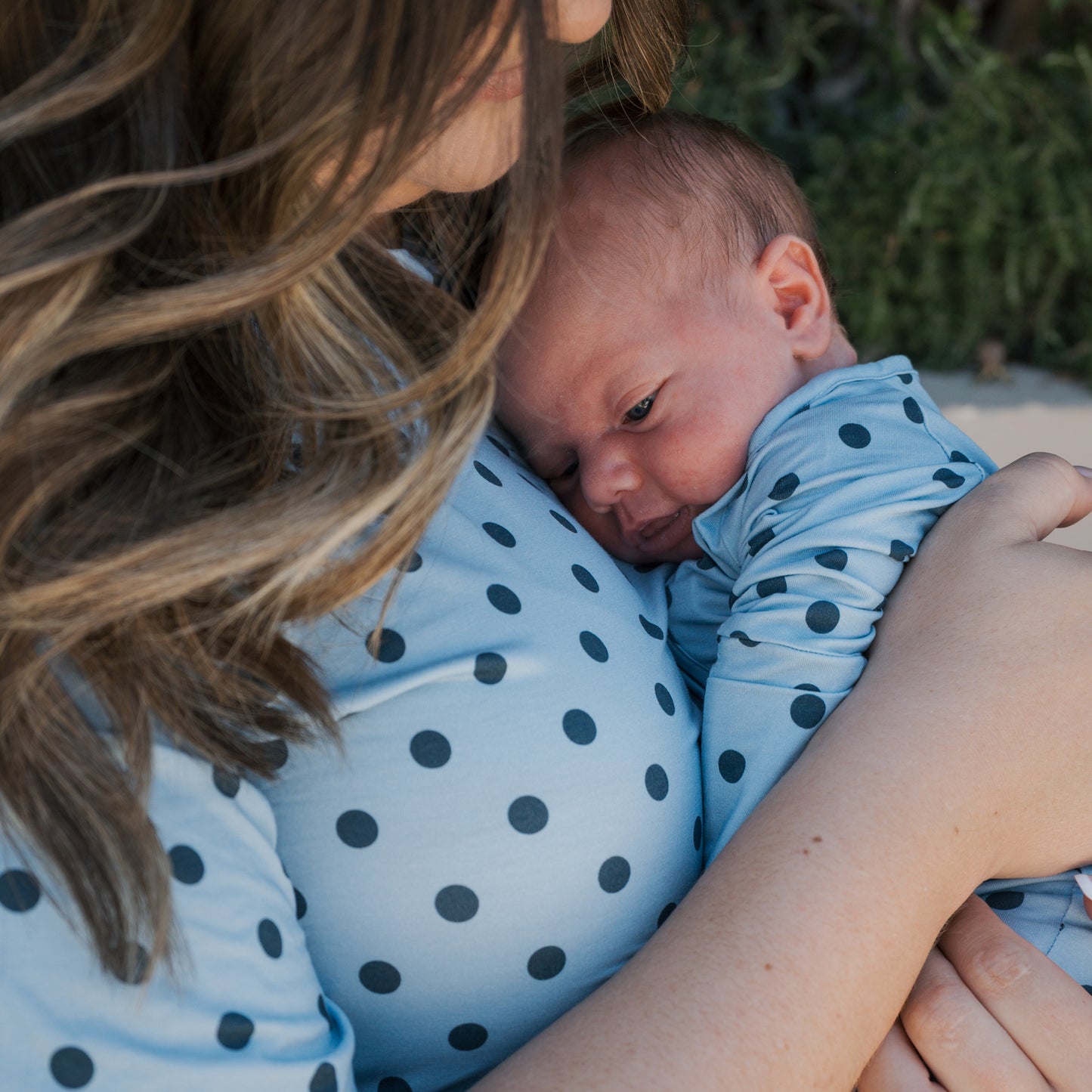 A woman with long brown hair holds a baby close to her chest, both dressed in matching light blue forever french baby Bamboo Zip Pajamas | Sully, featuring black polka dots and made from breathable fabric. The baby rests its head on the woman as they cuddle together.