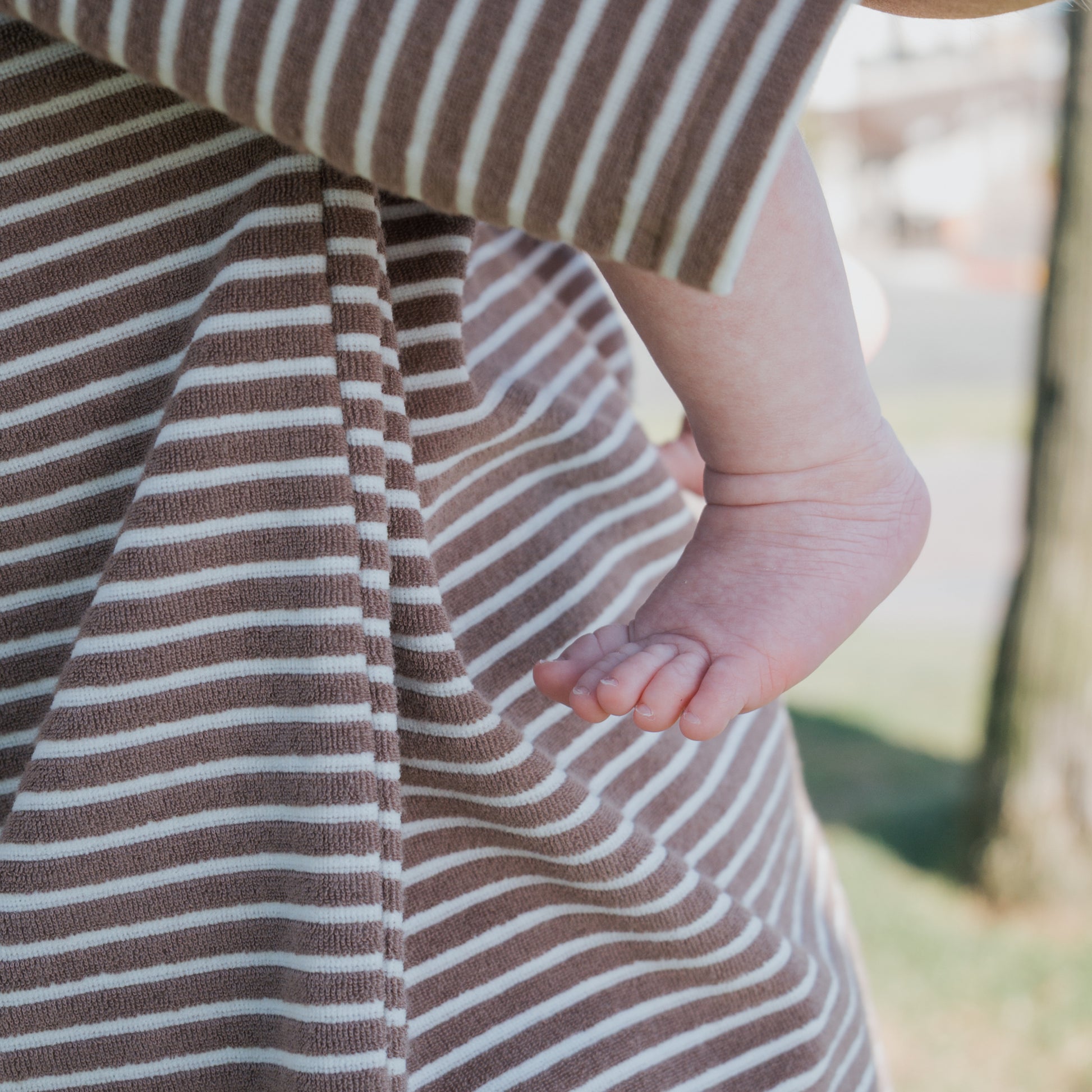 A close-up of a baby’s bare foot and leg, resting against an adult in stripes, highlights the forever french baby Terry T-Shirt Romper in Cola Stripe—ideal for coordinated family outfits with a soft, cozy touch.
