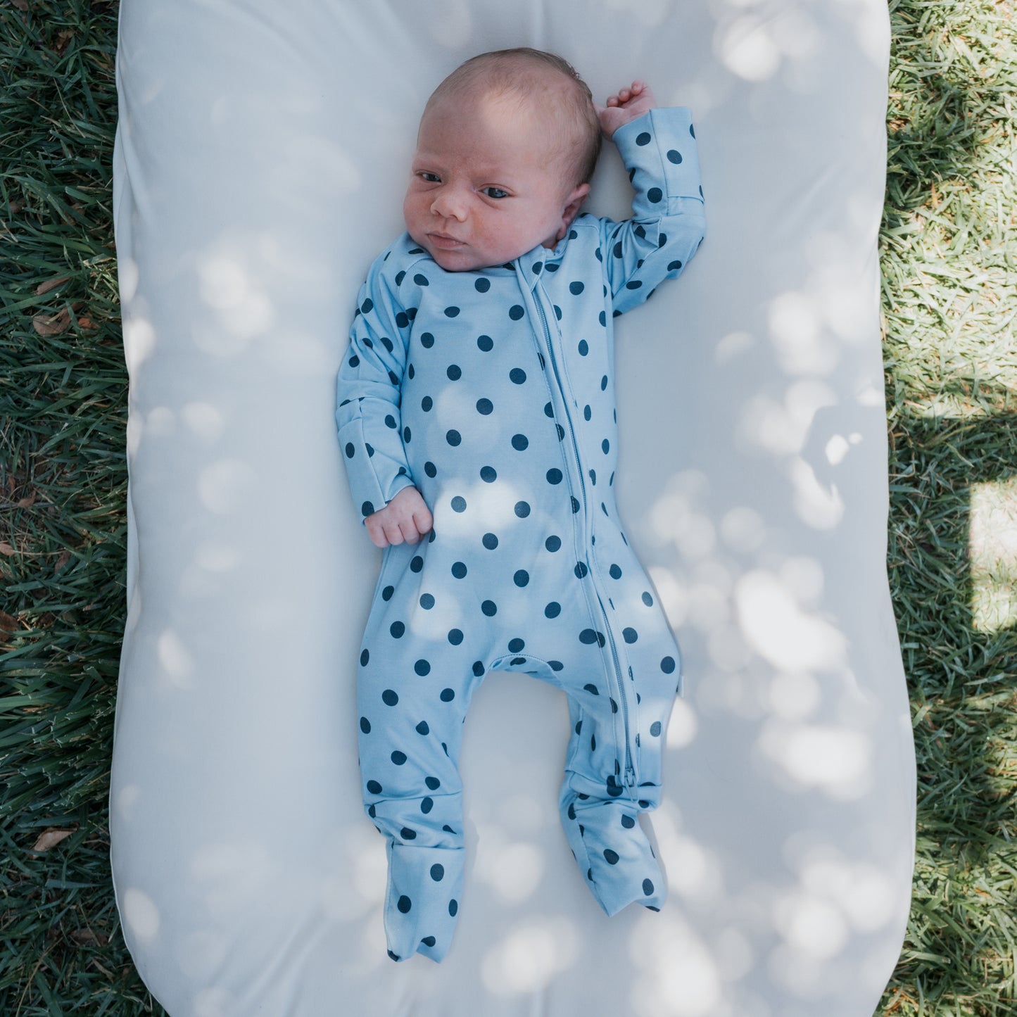 A baby in forever french baby’s Bamboo Zip Pajamas | Sully—featuring a blue polka-dot design and made from breathable bamboo fabric—lies on a white cushion outdoors, surrounded by grass and dappled sunlight.