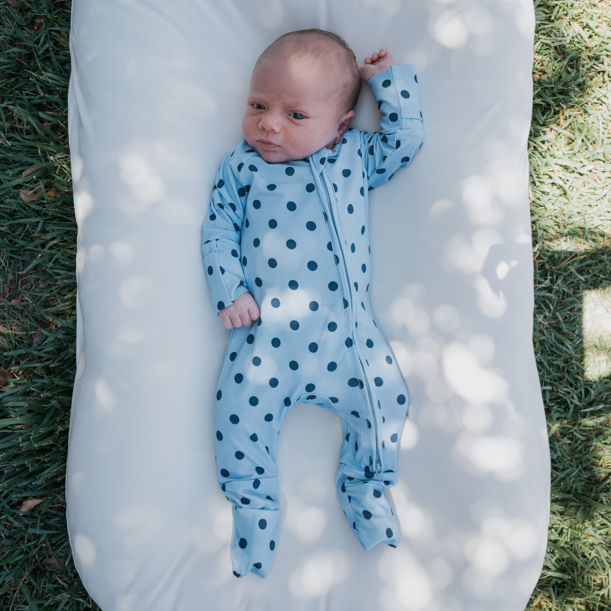 A baby in forever french baby’s Bamboo Zip Pajamas | Sully—featuring a blue polka-dot design and made from breathable bamboo fabric—lies on a white cushion outdoors, surrounded by grass and dappled sunlight.