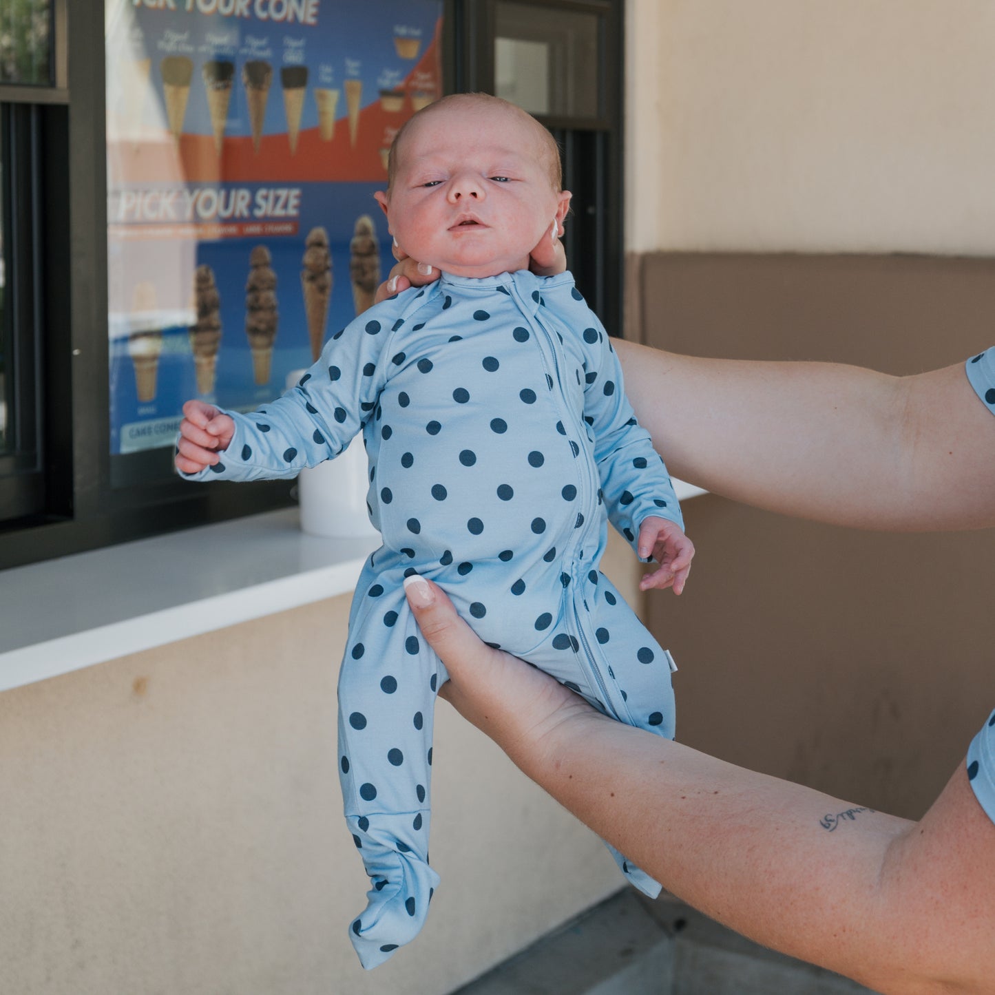 A baby wearing the forever french baby Bamboo Zip Pajamas | Sully, a breathable blue polka dot onesie, is held upright by an adult outside an ice cream shop, with a menu of ice cream cones visible in the background.