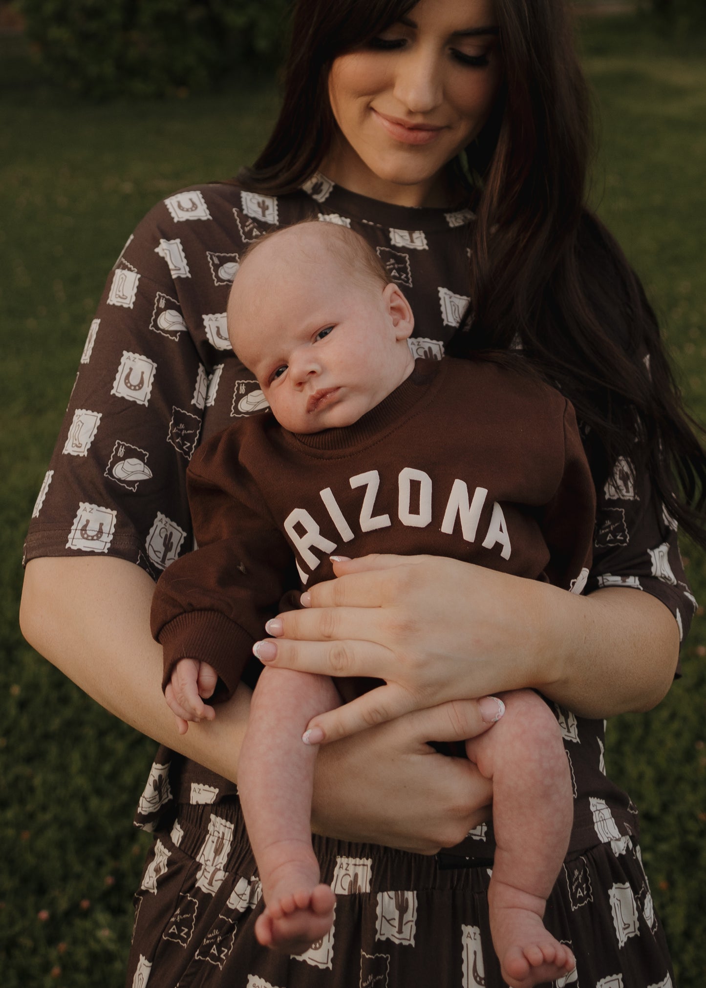 A woman in a patterned brown dress holds her baby in a forever french baby Sweatshirt Romper | ff Arizona. On the grass outdoors, she smiles gently, highlighting their matching outfits as she lovingly looks down at her baby.