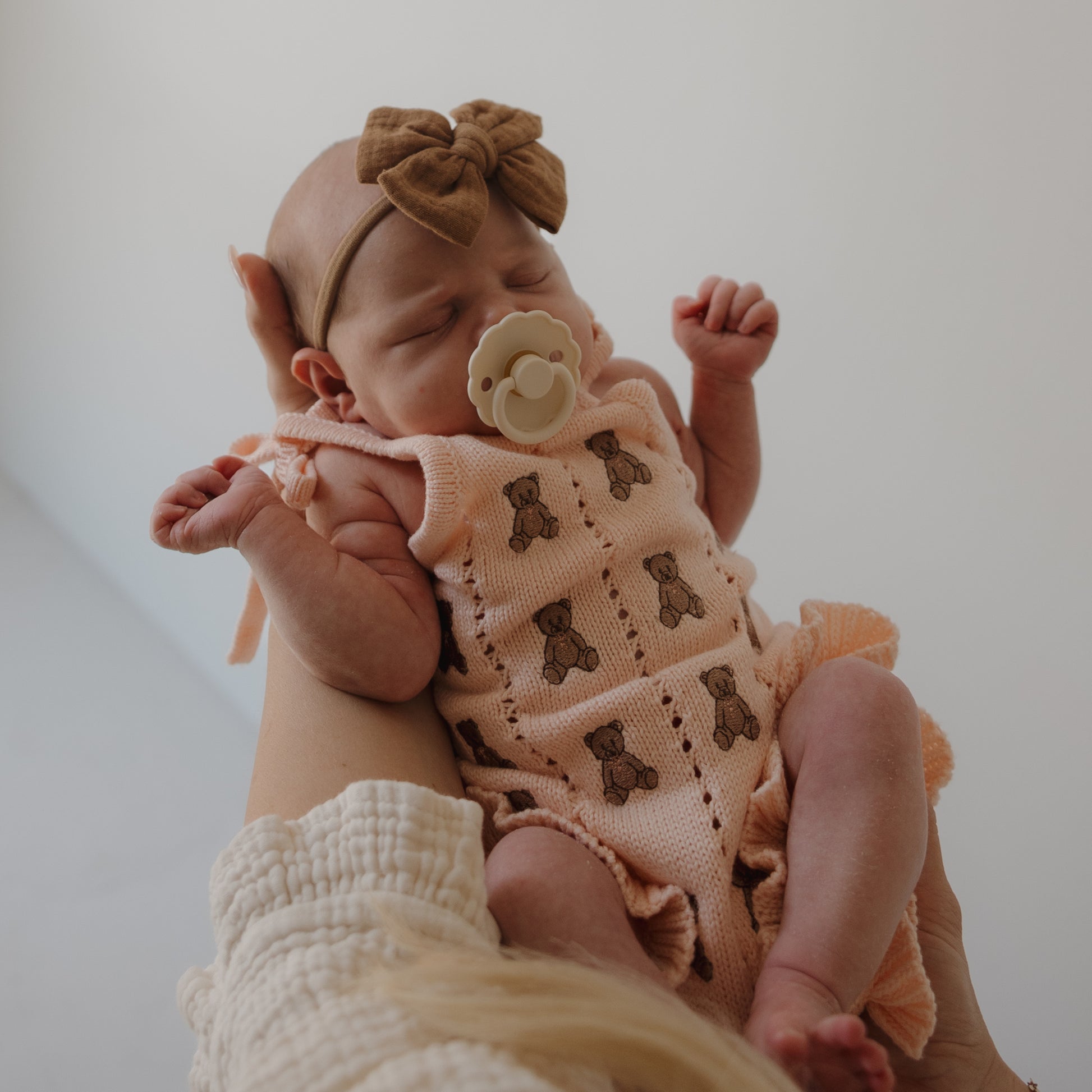 A baby girl in the forever french baby Knit Ruffle Romper | Lottie, featuring peach embroidery, ruffles, and a matching brown bow headband, sleeps peacefully with a pacifier while being gently held.
