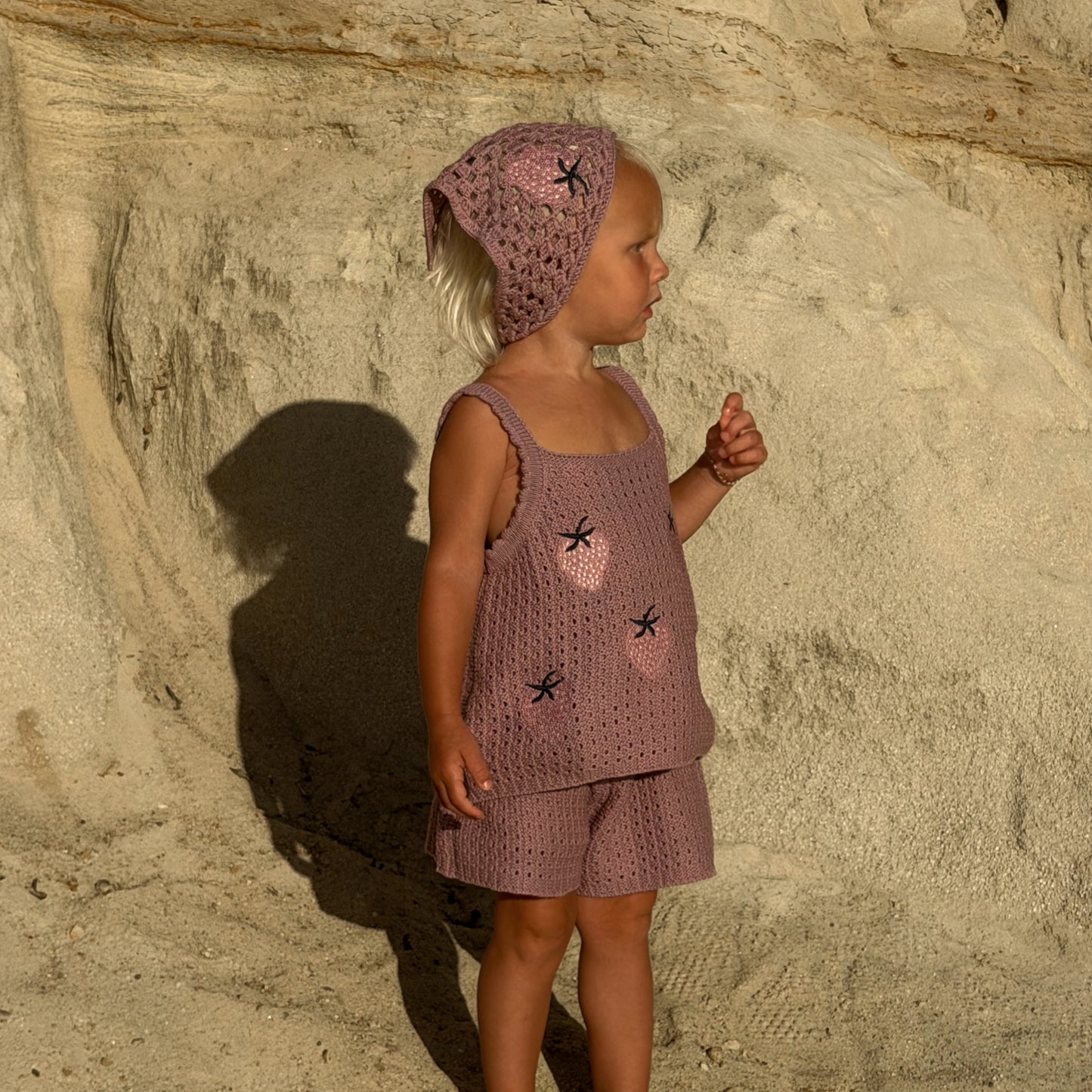 A young child stands sideways on sandy ground in warm sunlight, facing beige rock formations and wearing a matching pinkish crocheted top and shorts, along with the forever french baby Knit Head Scarf | Matilda, a cotton yarn accessory featuring black star accents and an adjustable tie.
