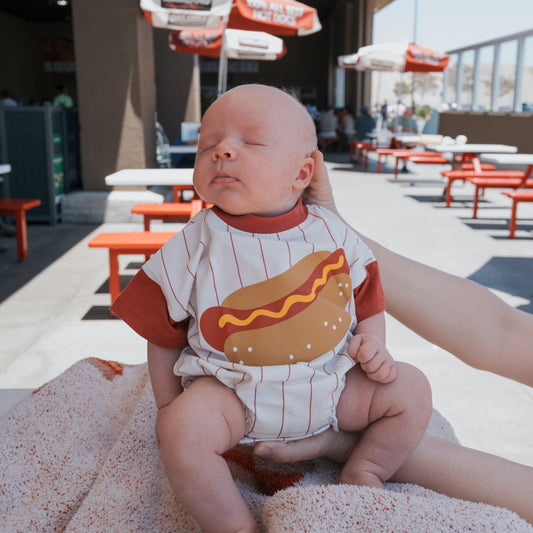 A baby wears the forever french baby Short Sleeve Romper | Hot Dog, sitting calmly with closed eyes as an adult supports them outdoors among red tables and umbrellas—a perfect moment for family matching outfits.