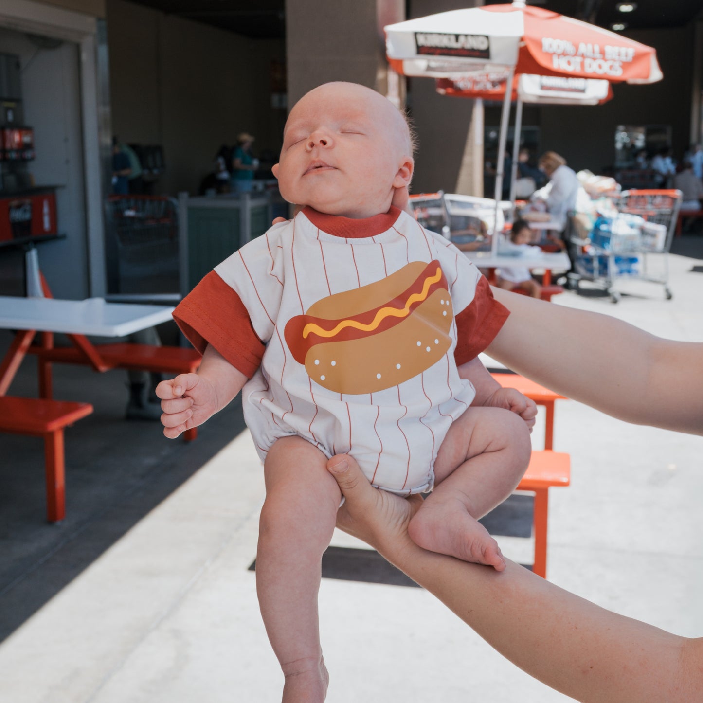 A baby wears the forever french baby Short Sleeve Romper | Hot Dog while being held outside a food court with red tables, chairs, and umbrellas on a sunny day.