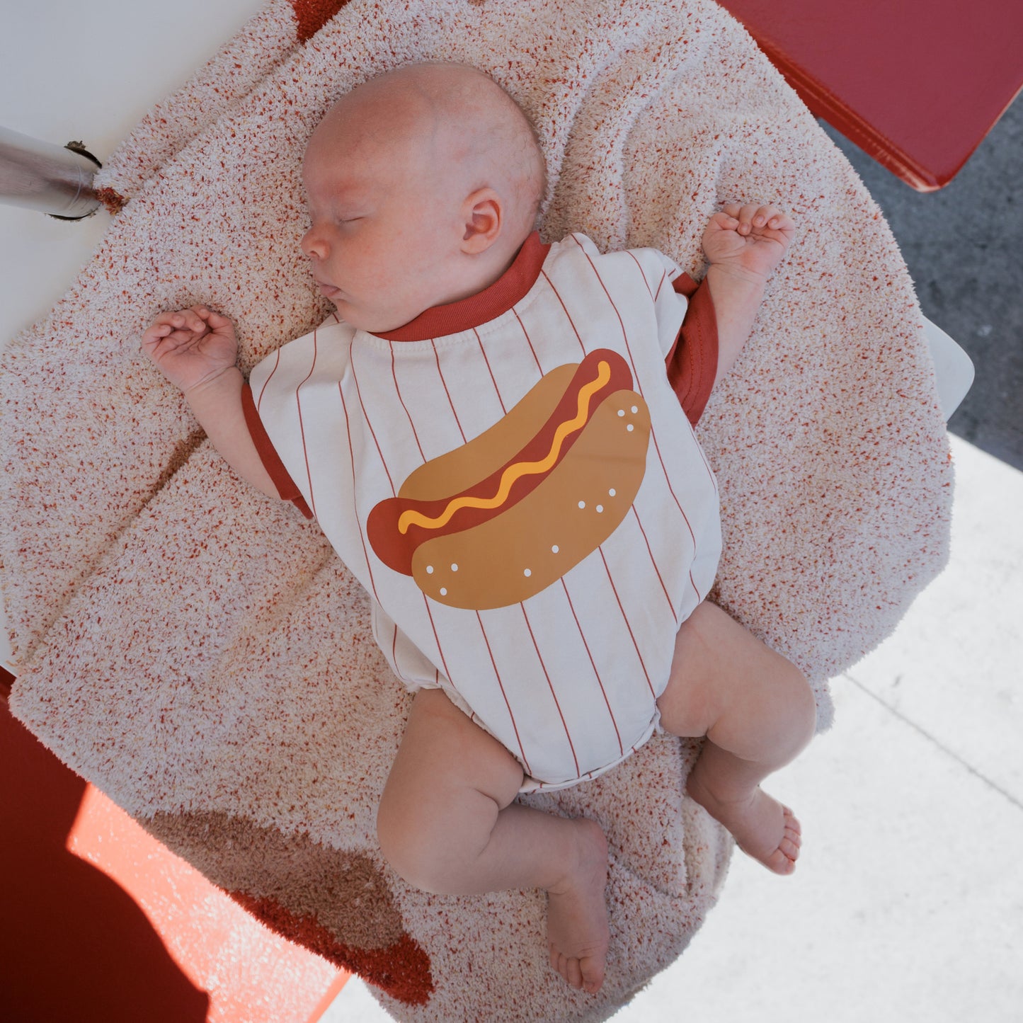 A baby naps on a textured blanket, wearing the forever french baby Short Sleeve Romper | Hot Dog—a cotton romper with red stripes and a big hot dog graphic. Bright daylight creates a cheerful vibe, ideal for matching family outfit photos.