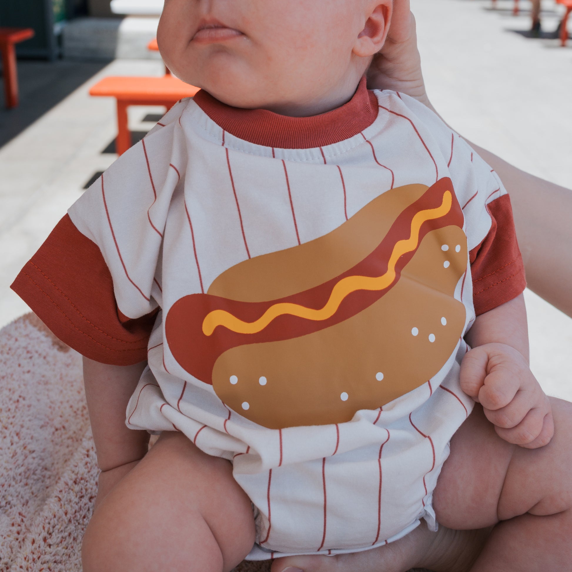 A baby wears the forever french baby Short Sleeve Romper | Hot Dog, a cute white and red cotton romper with a big hot dog graphic—bun, sausage, mustard—perfect for outdoor fun while being held by an adult.