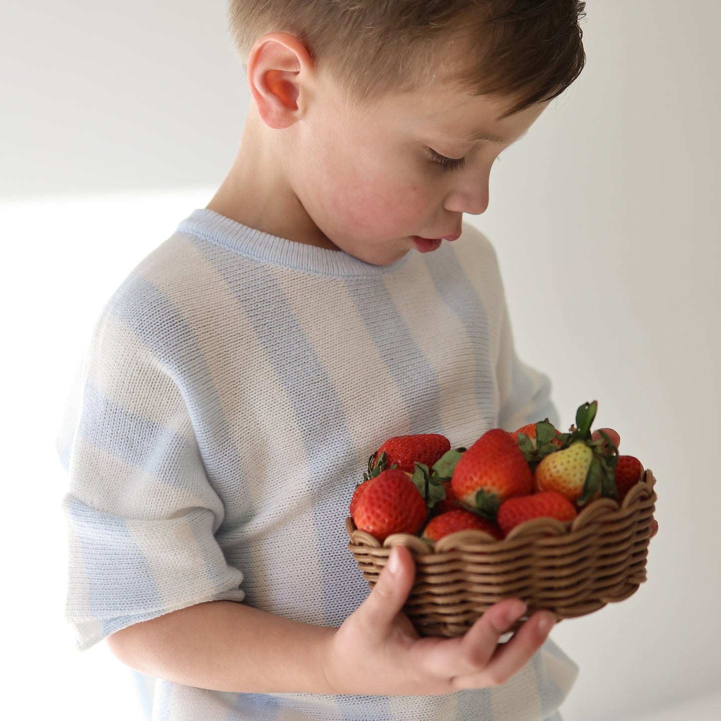 A young boy in the forever french baby Knit Short Set | Benny holds a wicker basket of fresh strawberries, looking down at the fruit against a softly lit, uncluttered background.