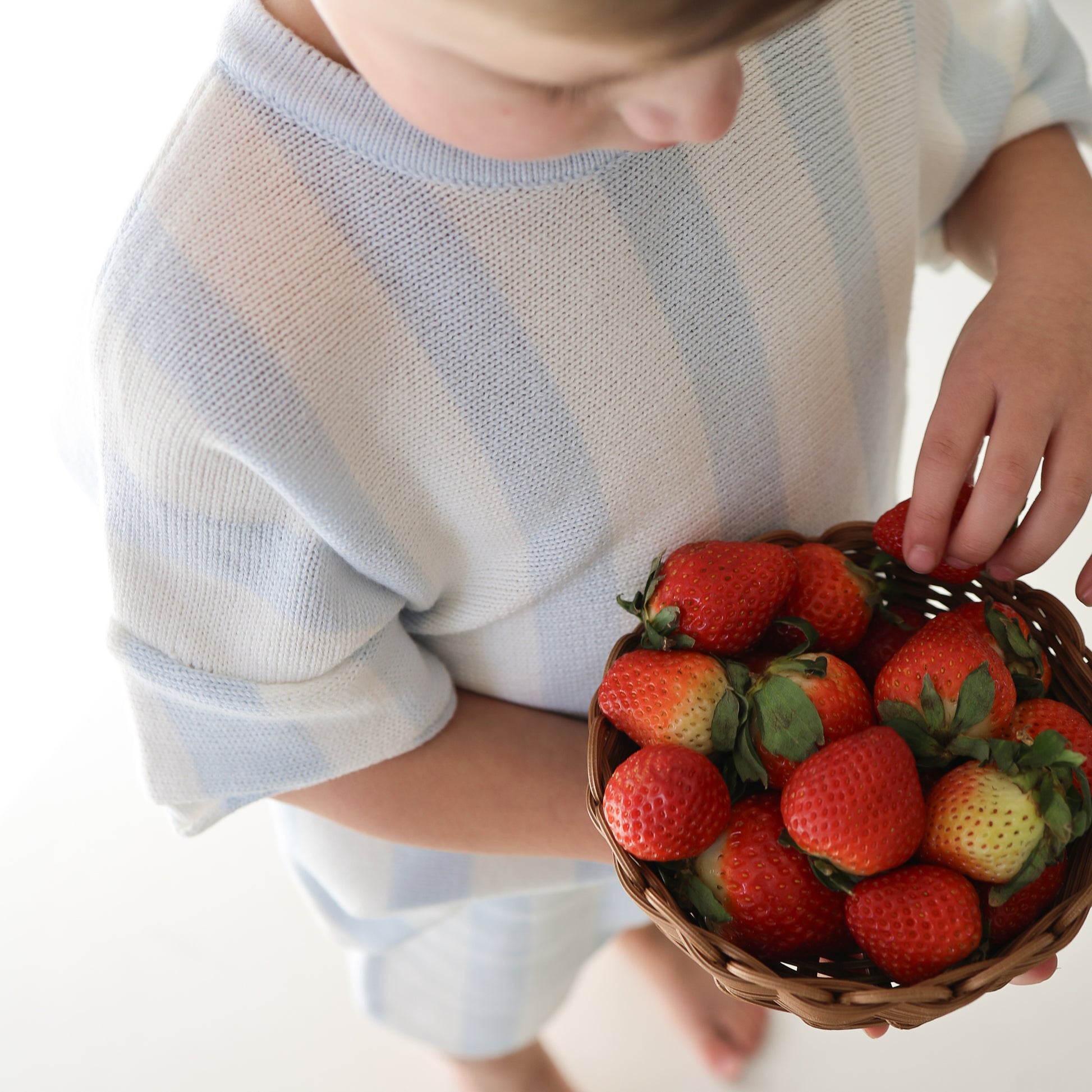 A child in a light blue and white Knit Short Set | Benny by forever french baby holds a basket of fresh strawberries, reaching in to pick one. The background is bright and minimal.