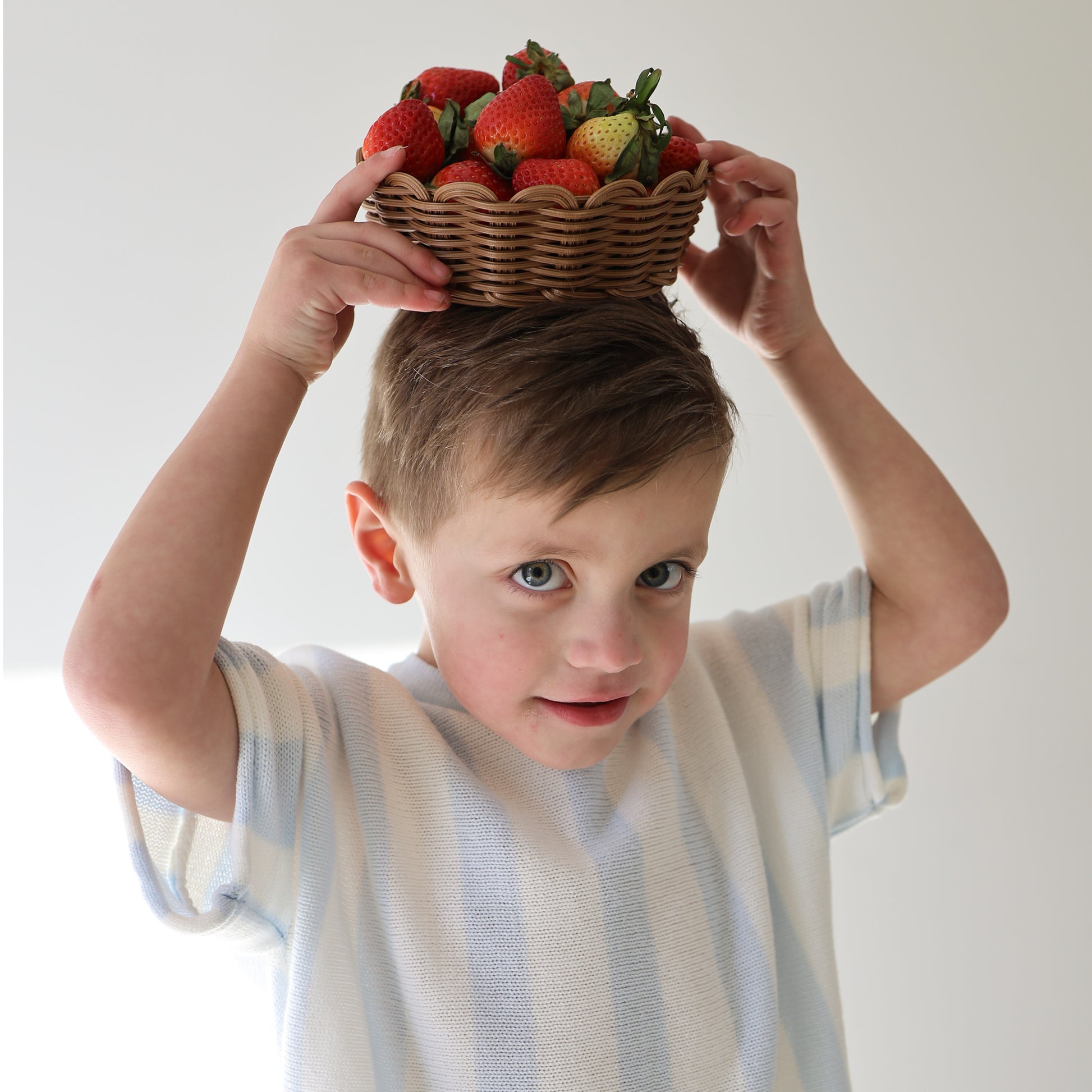 A young boy wears the forever french baby Knit Short Set | Benny and cabana stripe shorts, holding a wicker basket of strawberries on his head with a playful look, standing against a plain light background.