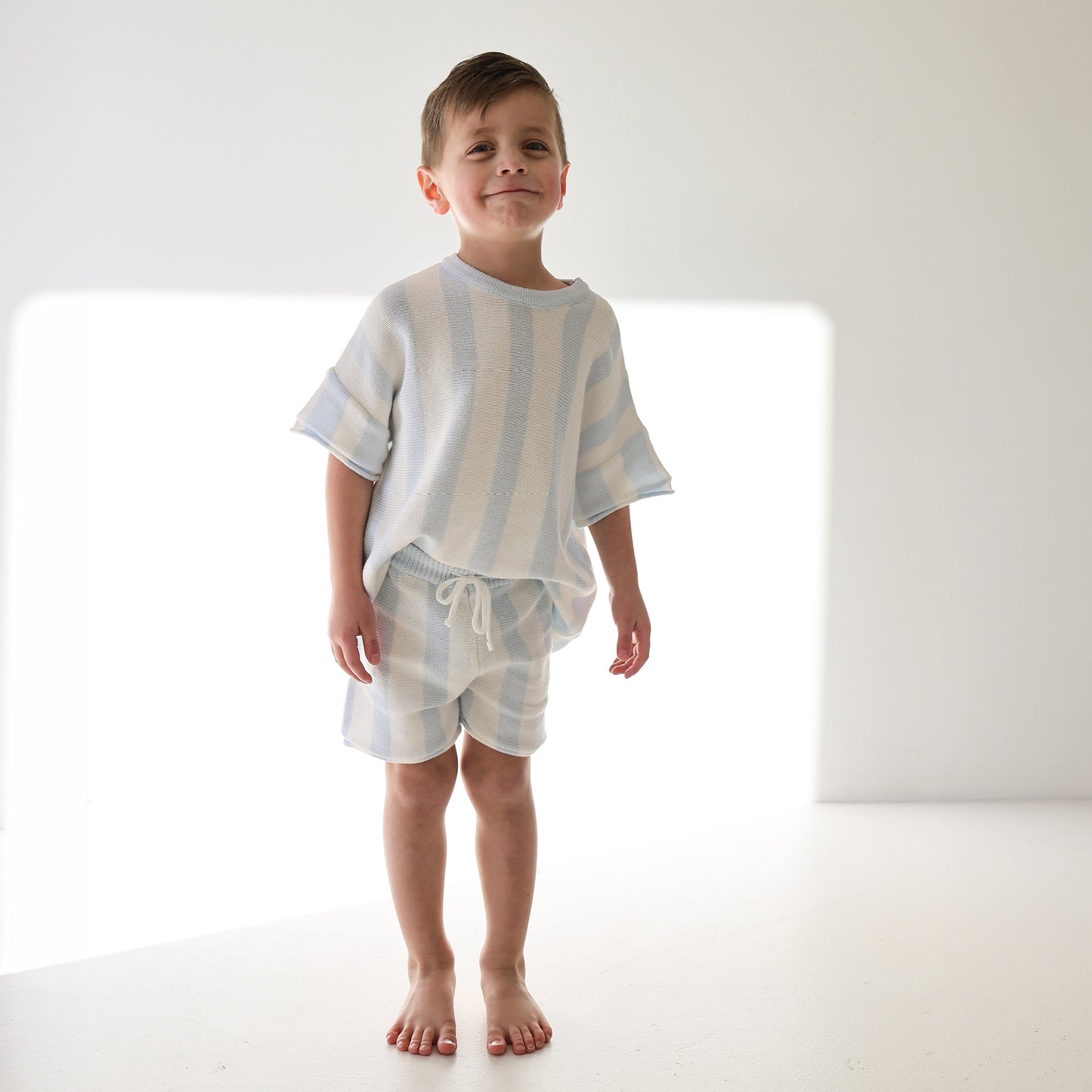 A young boy stands barefoot in a sunlit room, wearing the forever french baby Knit Short Set | Benny with light blue and white cabana stripe shorts. He smiles gently, hands resting by his sides.