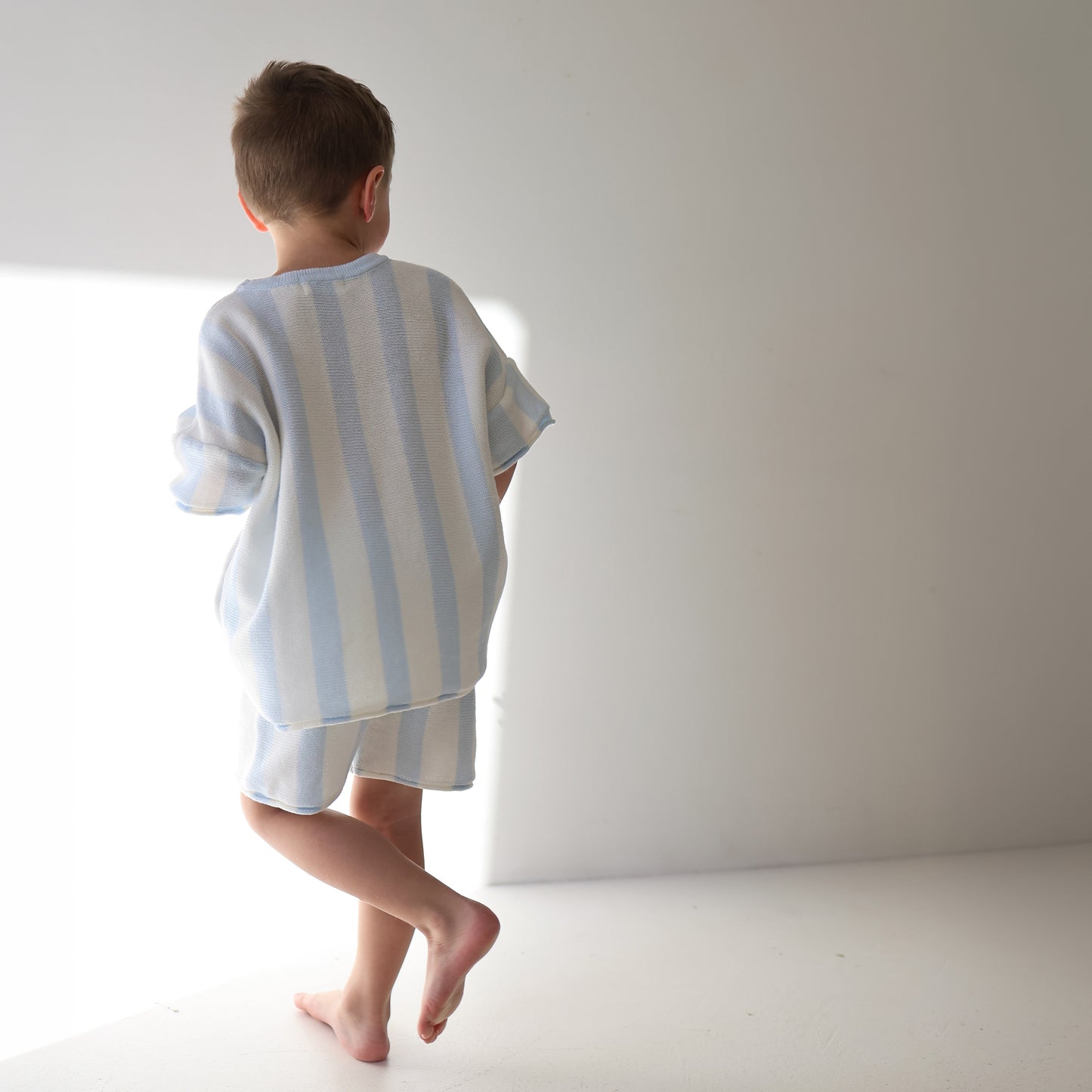 A young child stands barefoot in soft sunlight, facing away, wearing the forever french baby Knit Short Set | Benny—100% cotton with light blue and white cabana stripes—in a minimalist white room.