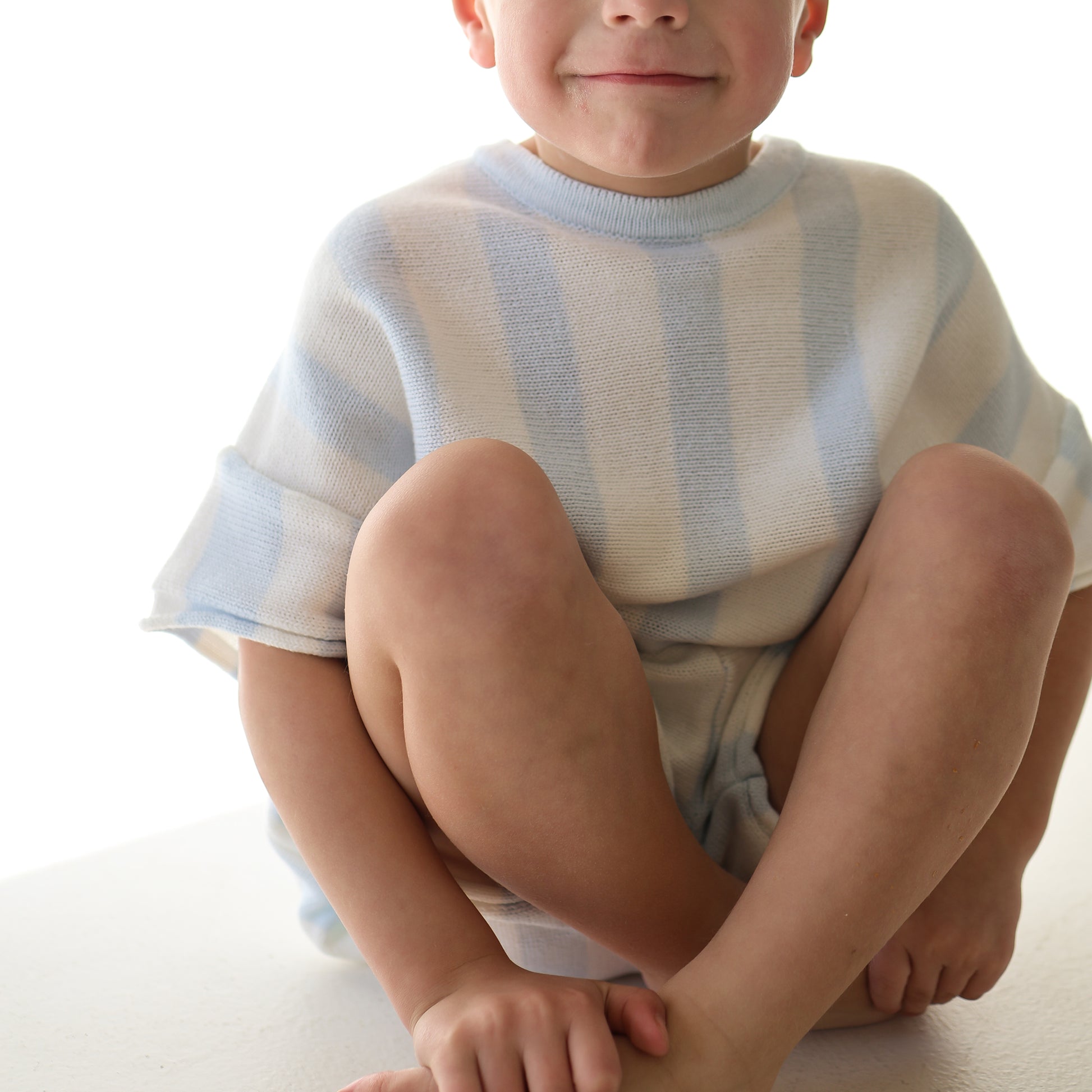 A young child smiles softly against a white background, wearing the forever french baby Knit Short Set | Benny—a comfy kids’ outfit in soft 100% cotton knit with light blue and white stripes.