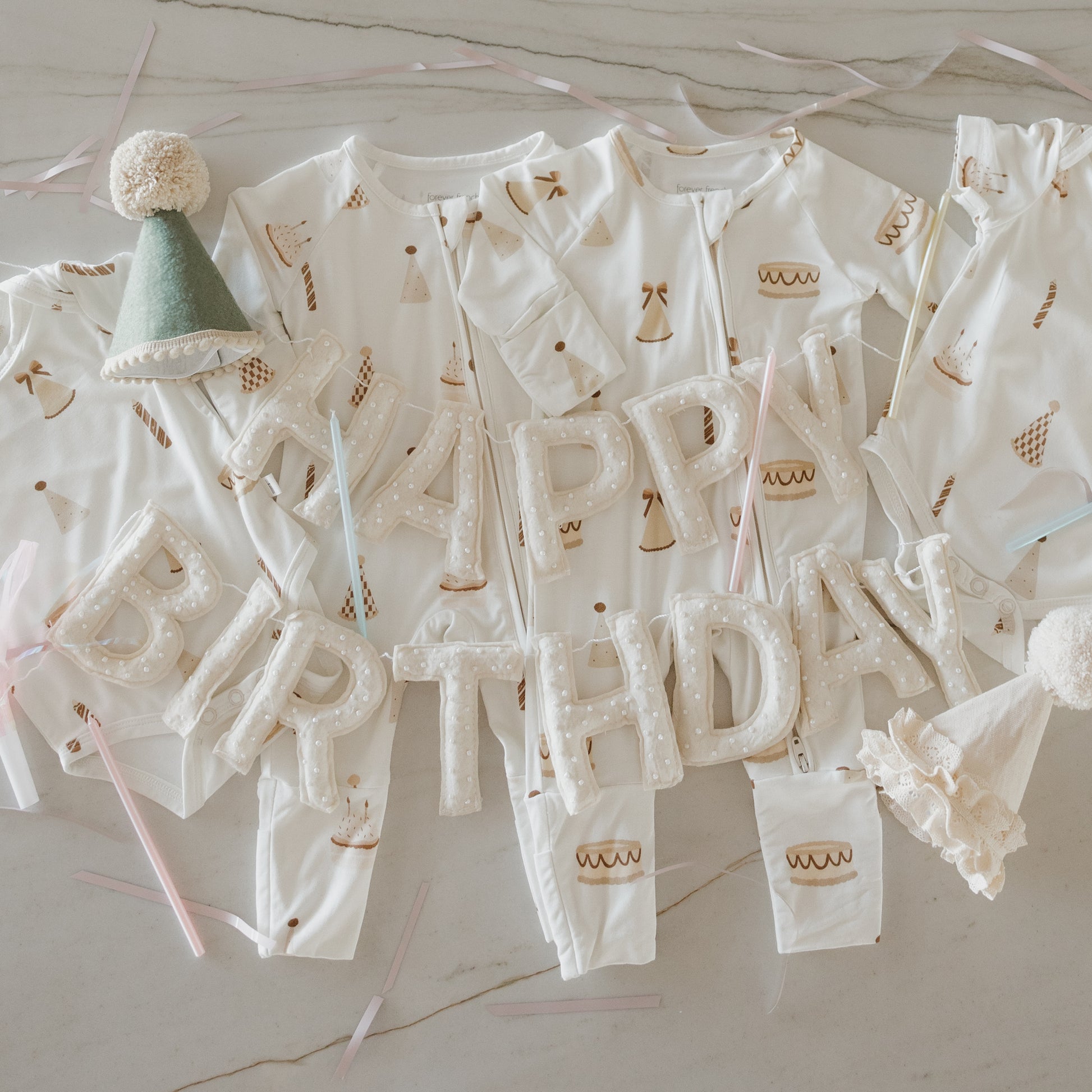 Forever French Baby’s Short Sleeve Bamboo Snapsuit | Birthday Boy, featuring charming cake prints, party hats, and balloon letters spelling “HAPPY BIRTHDAY,” is beautifully arranged on a light surface with pastel ribbons for an extra-sweet celebration. Crafted from hypoallergenic, breathable bamboo fabric, this snapsuit keeps your little one comfortable and adorable throughout their special day.