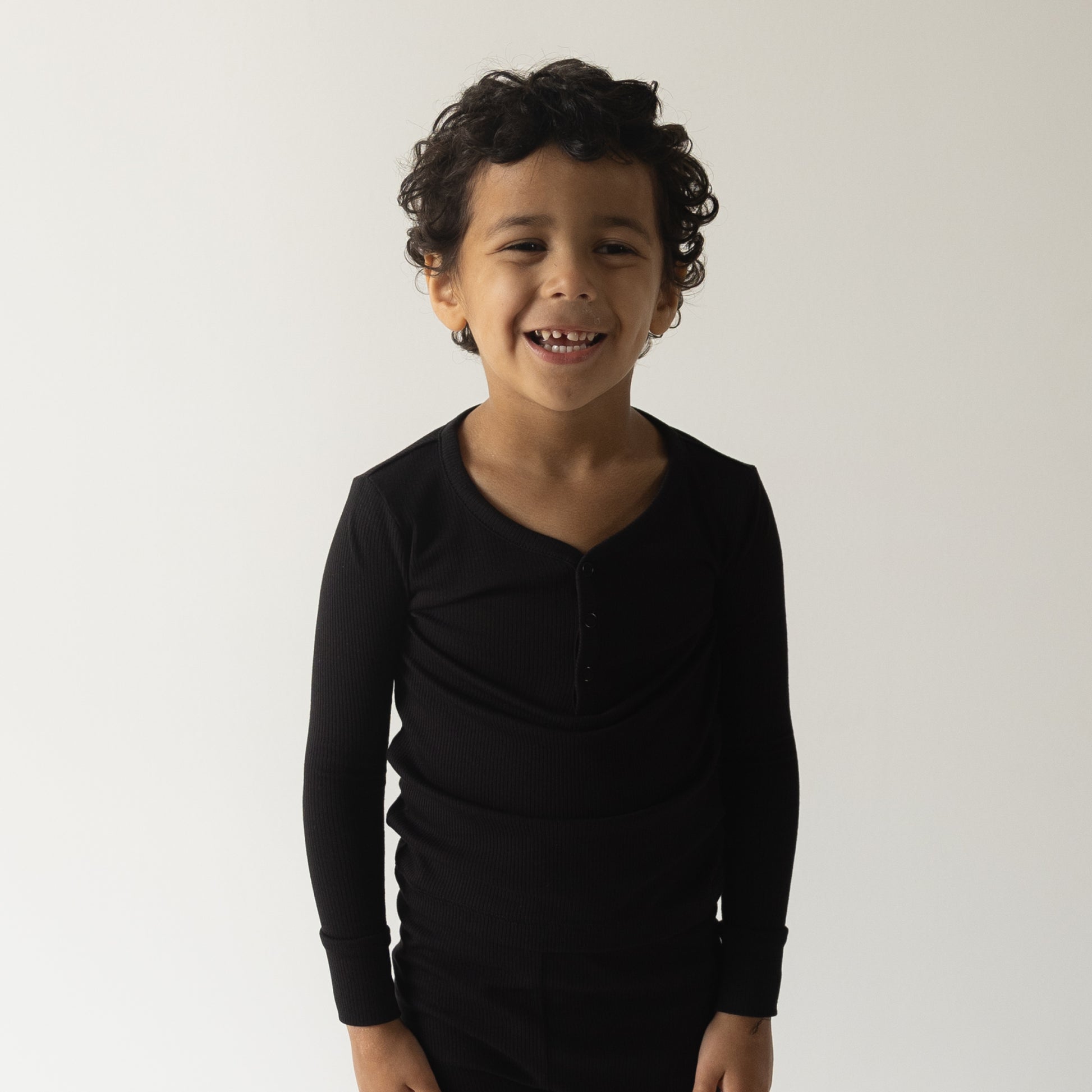 A young child with curly hair, wearing forever french baby’s Bamboo Two Piece Pajamas in Ribbed Black, smiles against a plain white background.