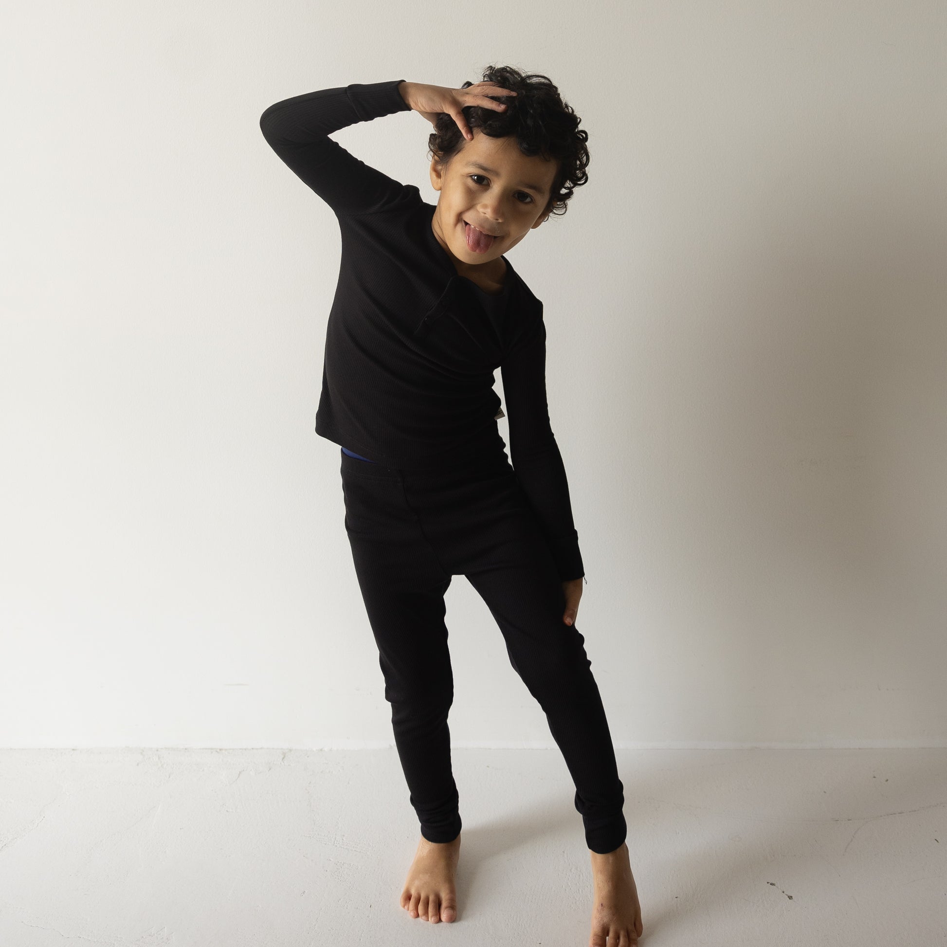 A child wearing forever french baby Bamboo Two Piece Pajamas | Ribbed Black stands barefoot on a white floor, smiling and playfully sticking out their tongue with one hand on their head against a plain white background.
