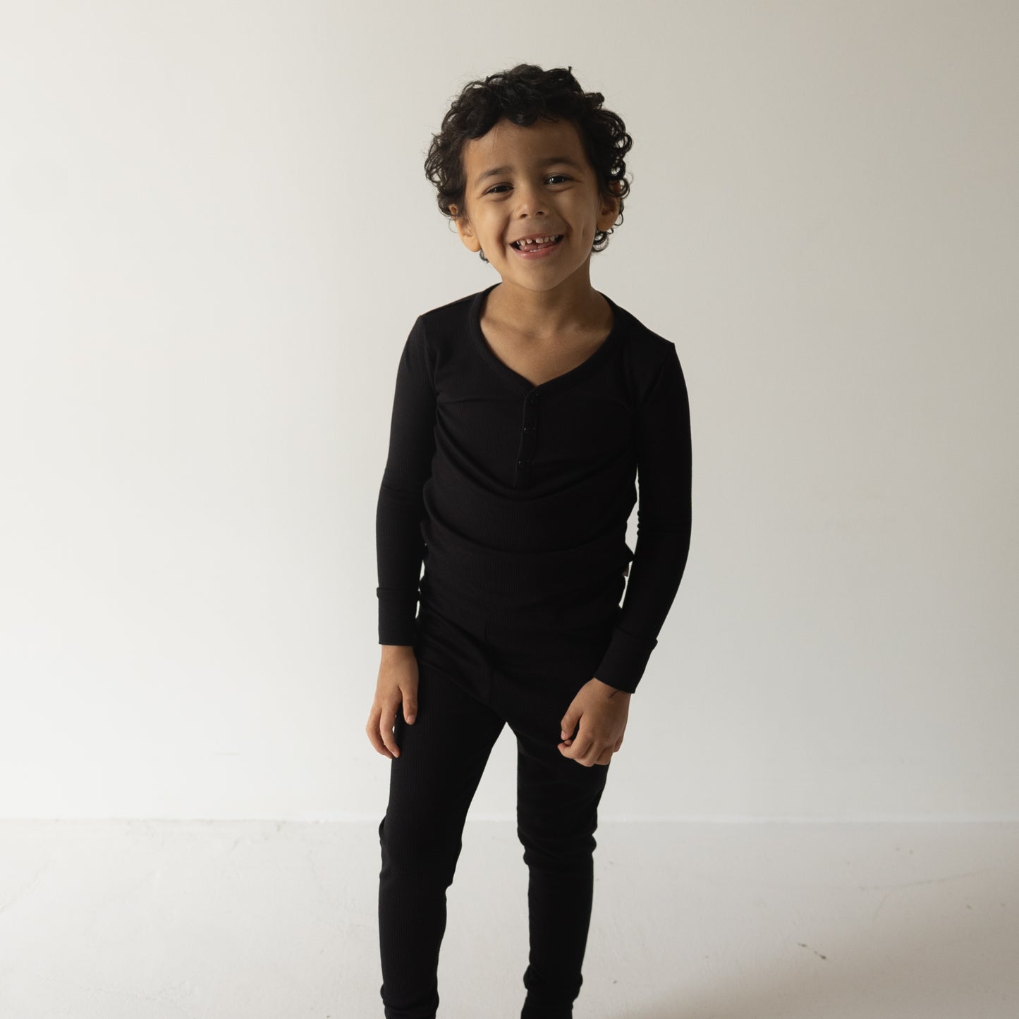 A young child with curly hair smiles against a white background, wearing forever french baby Bamboo Two Piece Pajamas in Ribbed Black—soft, hypoallergenic sleepwear ideal for sensitive skin.