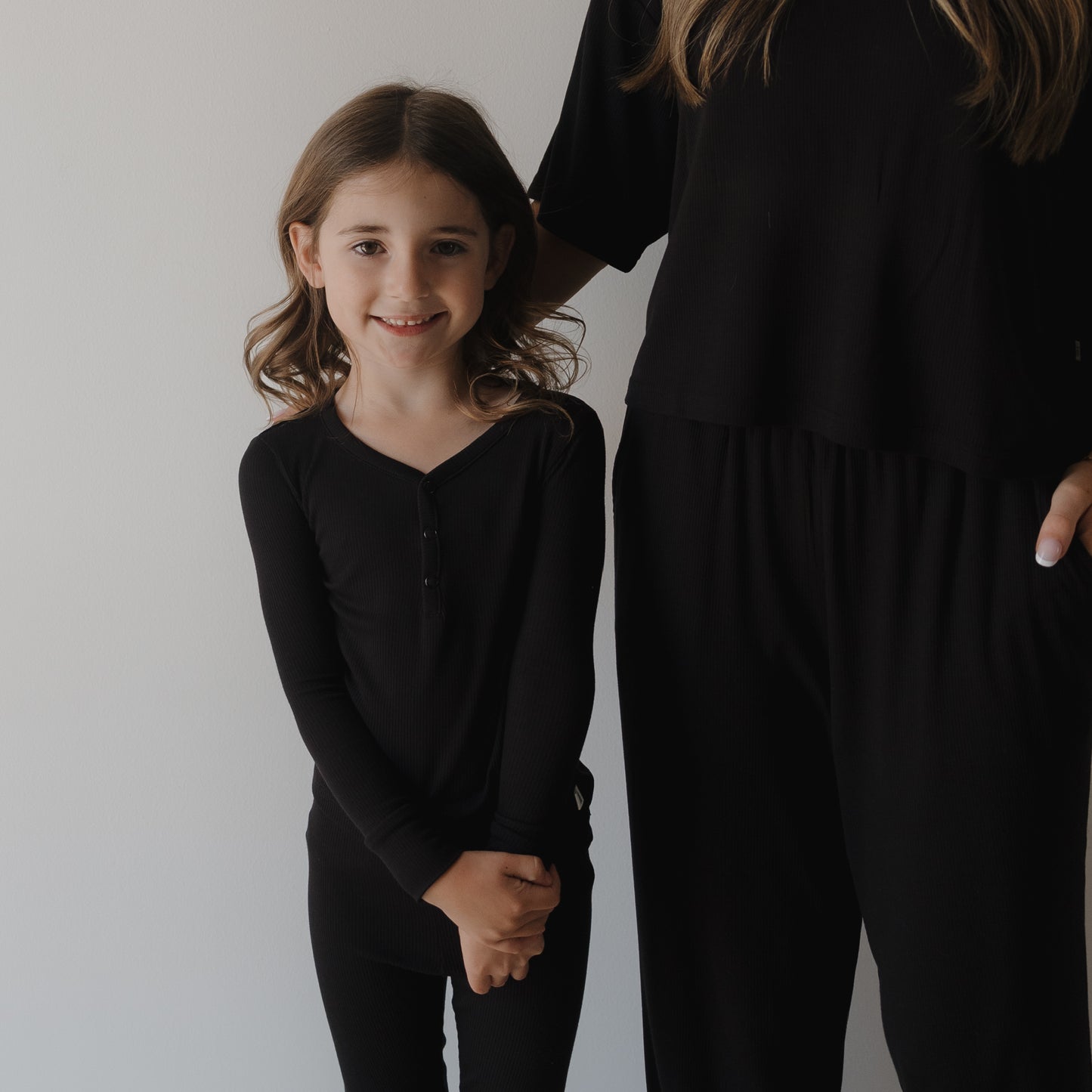 A young girl with wavy hair, smiling in forever french baby Bamboo Two Piece Pajamas | Ribbed Black, stands next to a partially visible adult in loose black clothing against a plain light background.