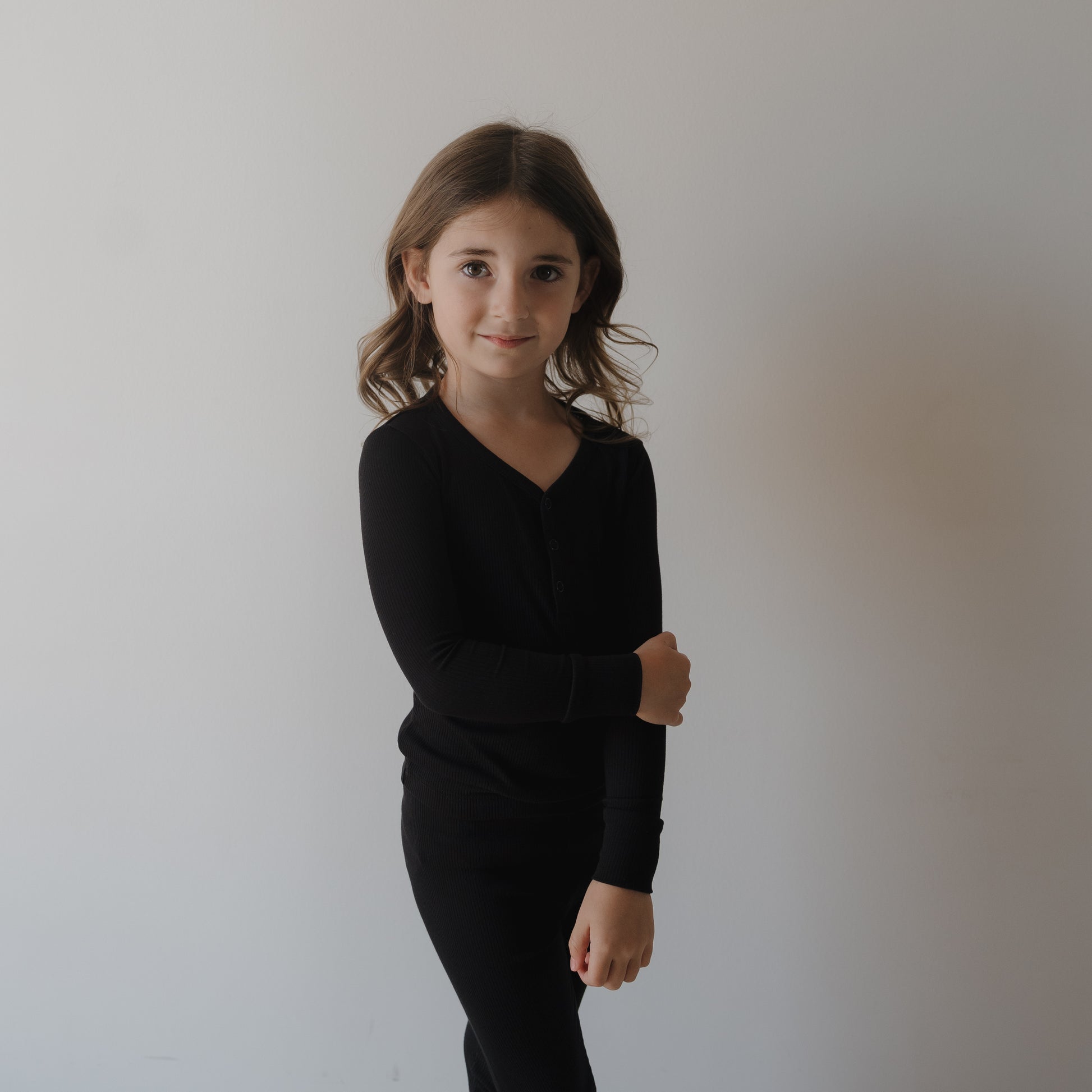 A young girl with wavy brown hair stands against a white wall, gently holding her arm and smiling softly. She wears the forever french baby Bamboo Two Piece Pajamas | Ribbed Black, made from hypo-allergenic fabric.